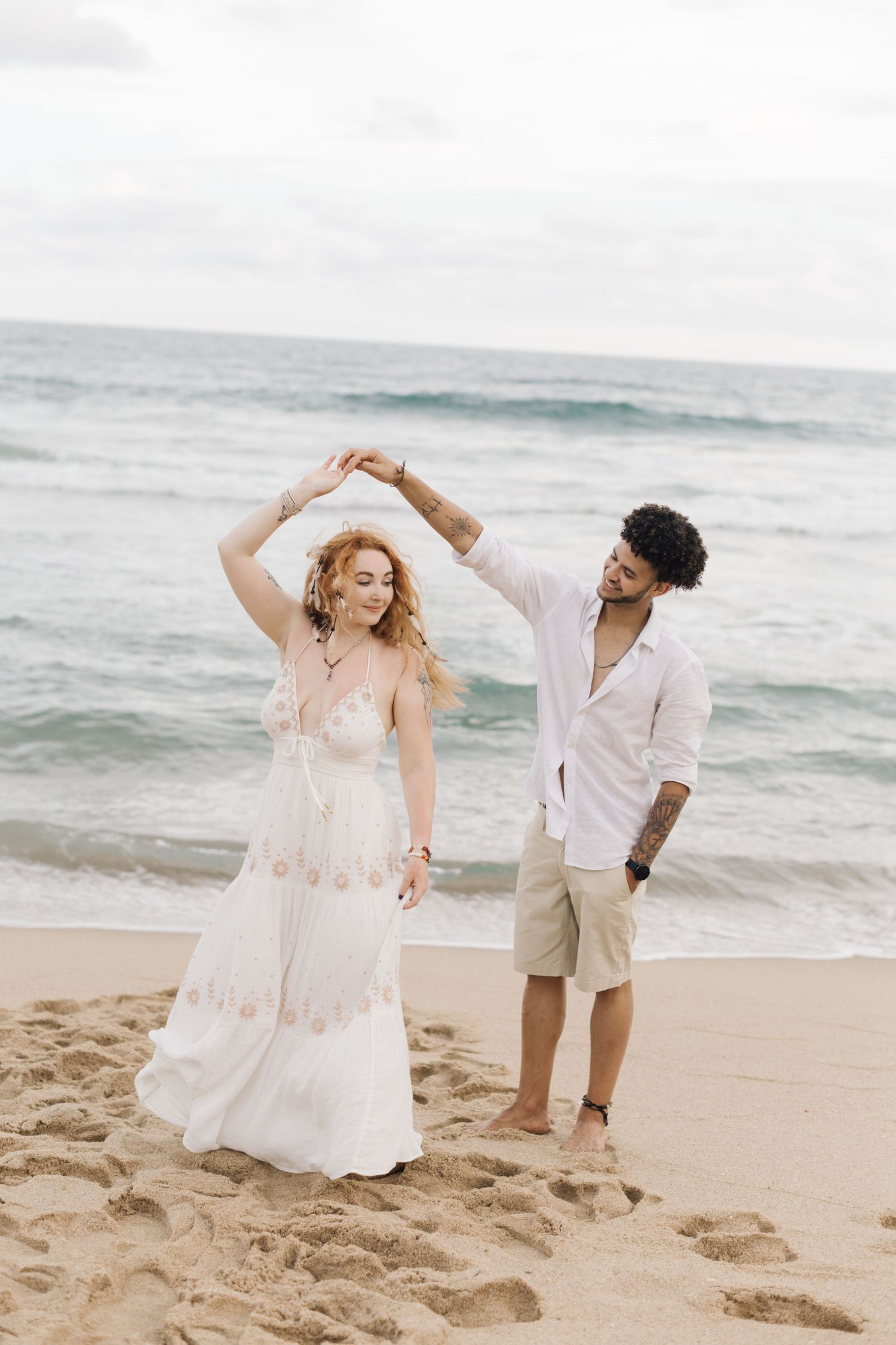 A couple in white clothing dances together on a beach with ocean waves in the background.