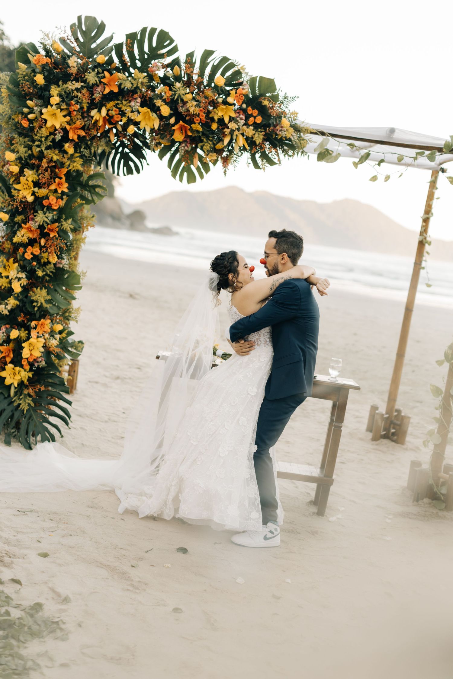 Wedding couple shares intimate moment under orange floral arch on sandy beach at sunset.