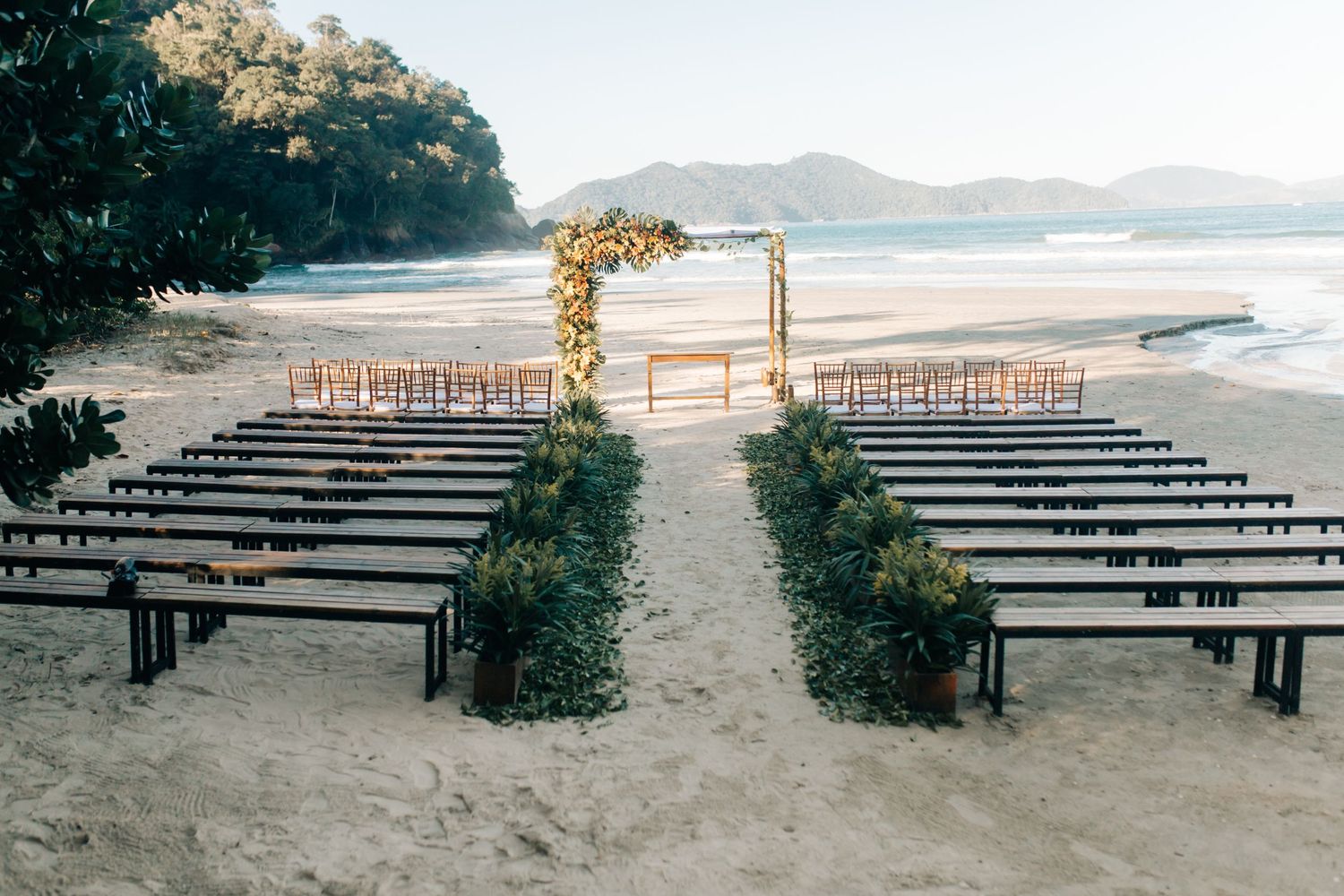 Beach wedding ceremony setup with wooden benches and floral arch facing ocean view.