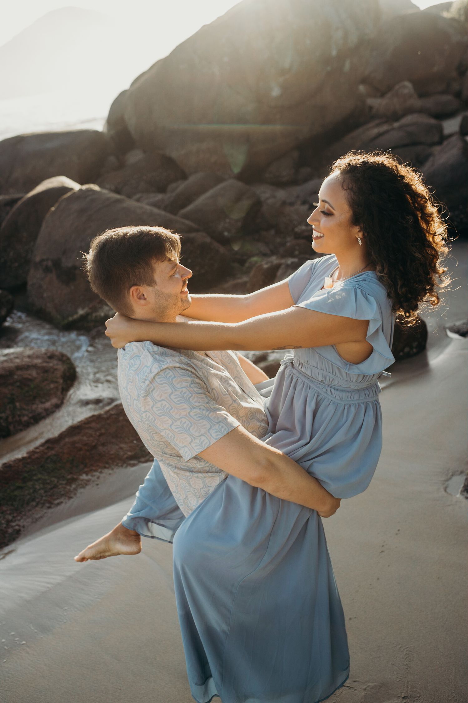 Playful couple dancing and laughing together on a rocky beach at sunset.