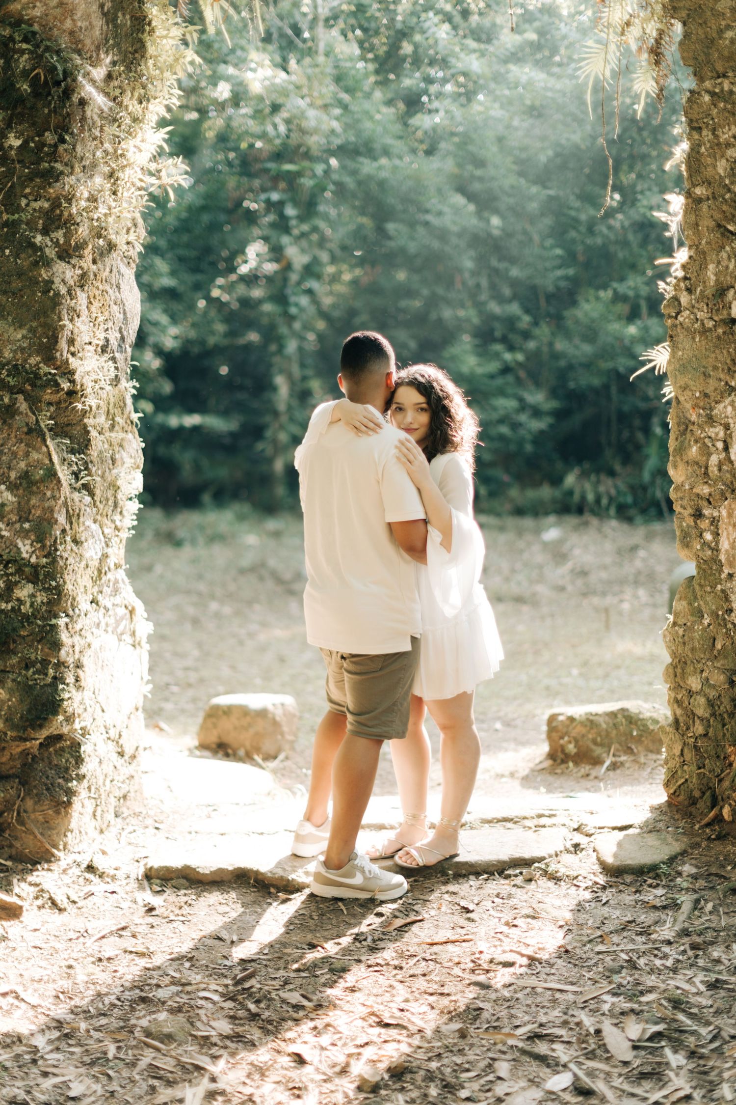 A romantic couple embraces in a sunlit stone archway surrounded by lush greenery.