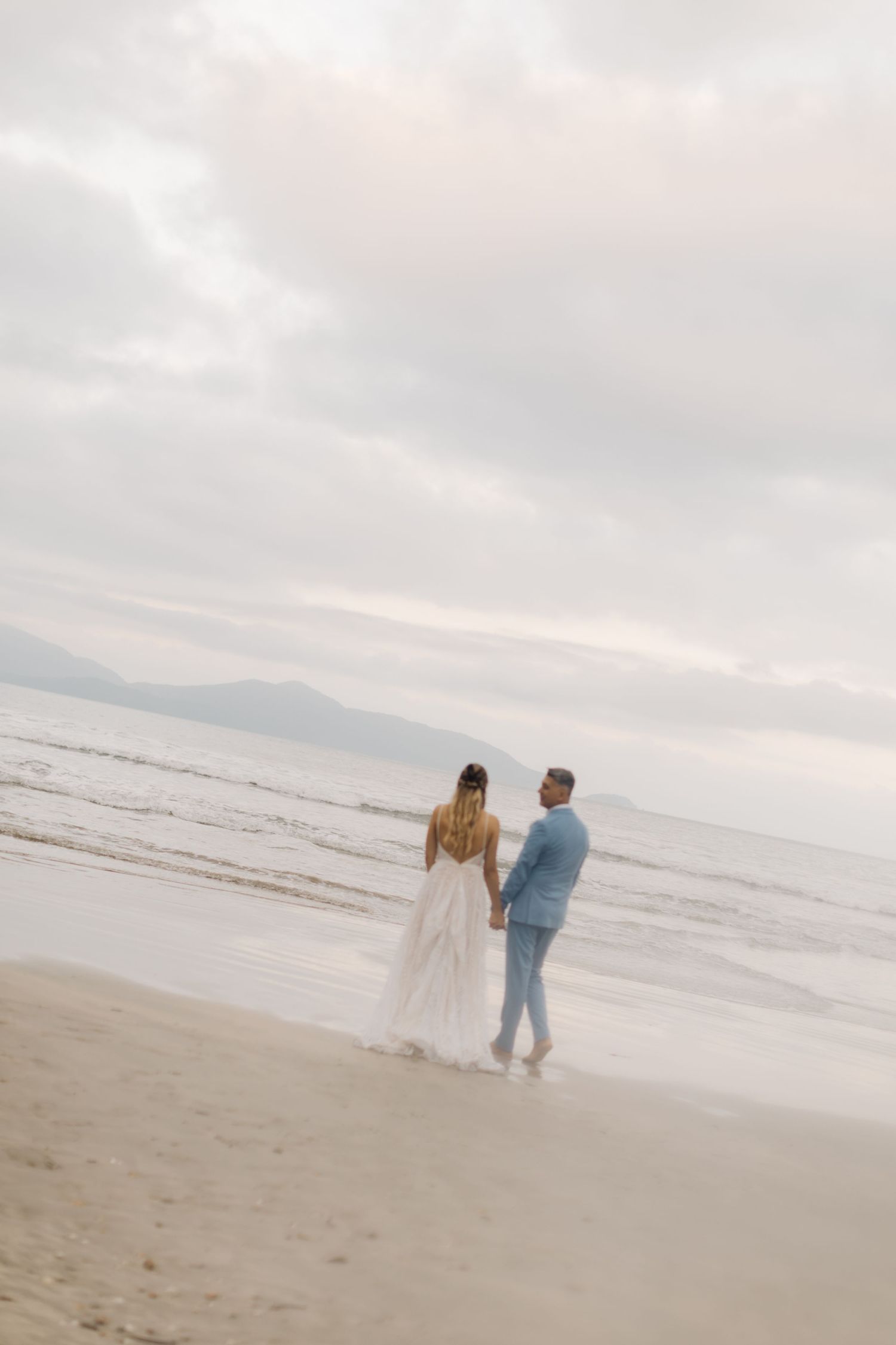 Couple in wedding attire standing on a misty beach at sunset looking out at the ocean and mountains.