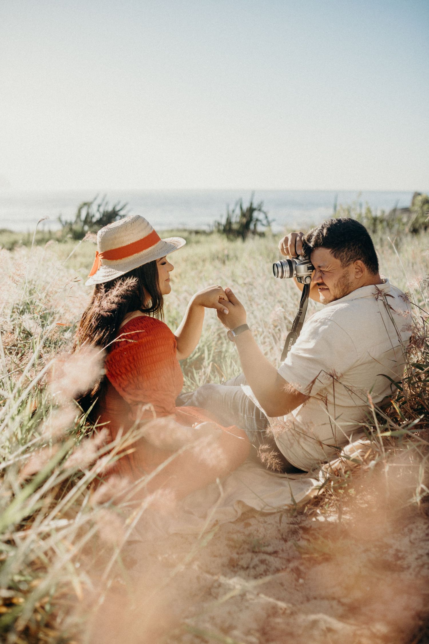 Two people hold hands while relaxing in a field as one photographs the coastal scenery.