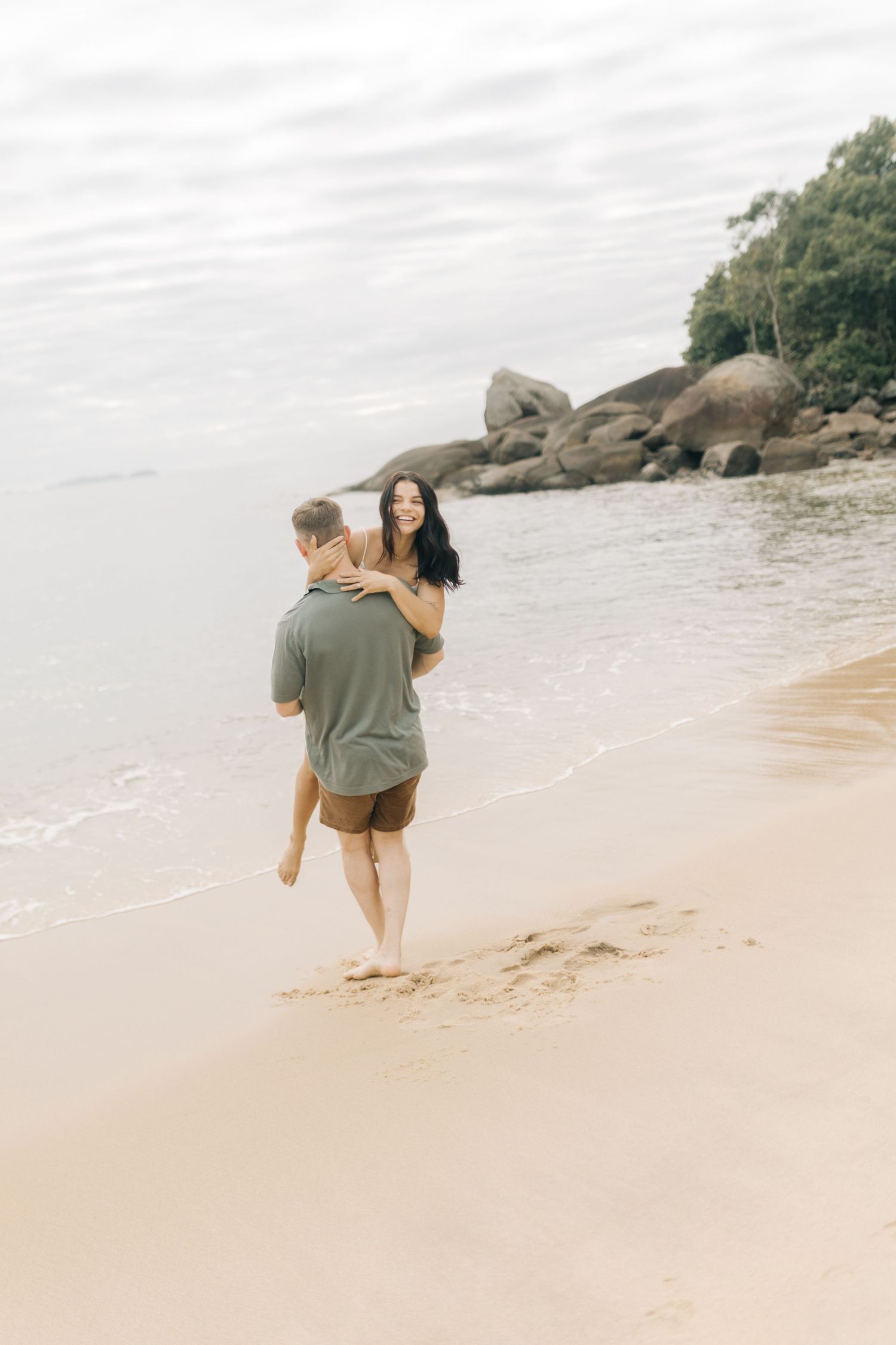 A tender beach moment between a couple walking along the shoreline near large coastal rocks.
