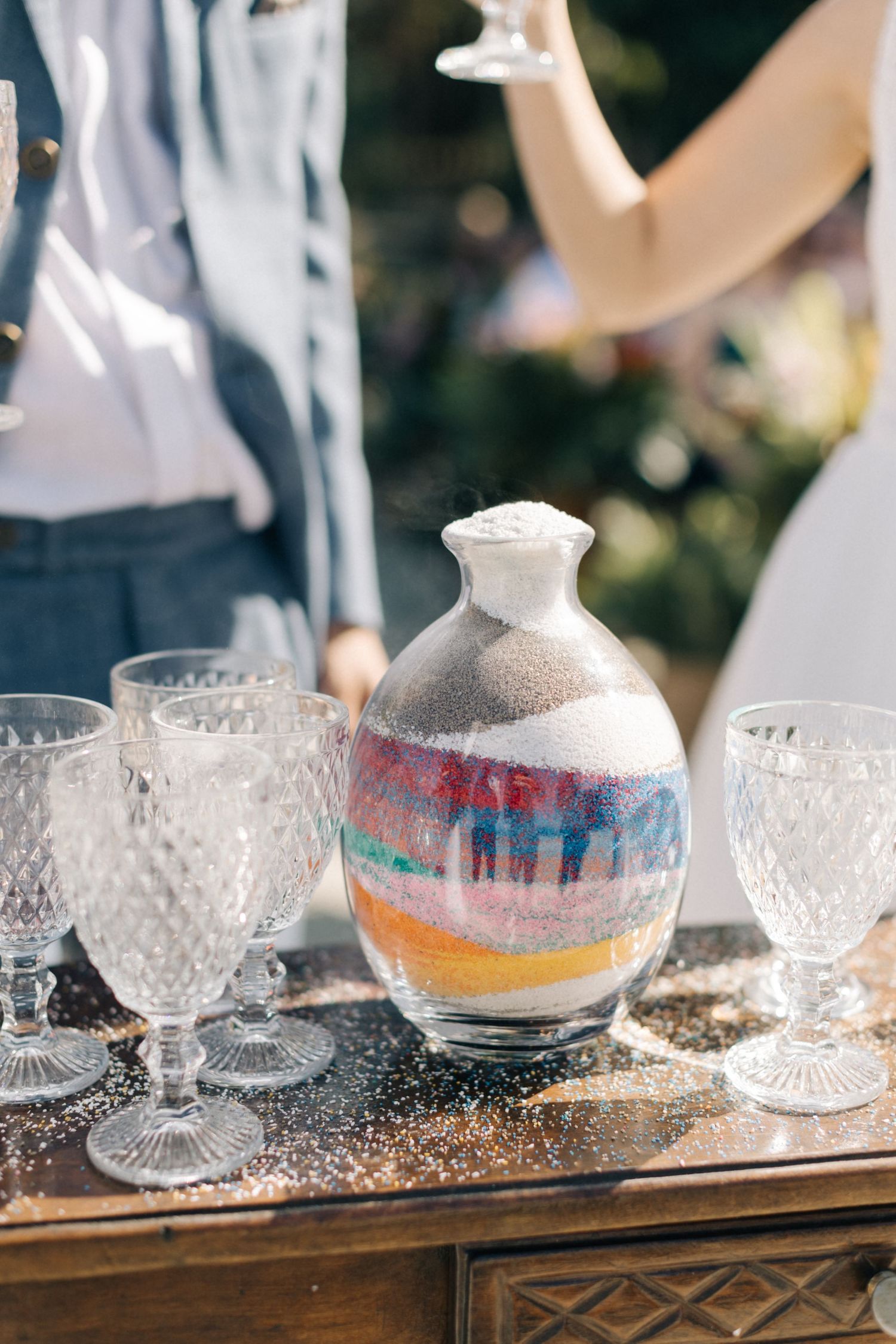 Decorative glass vessel with colorful layered sand surrounded by crystal glasses on a wooden table.