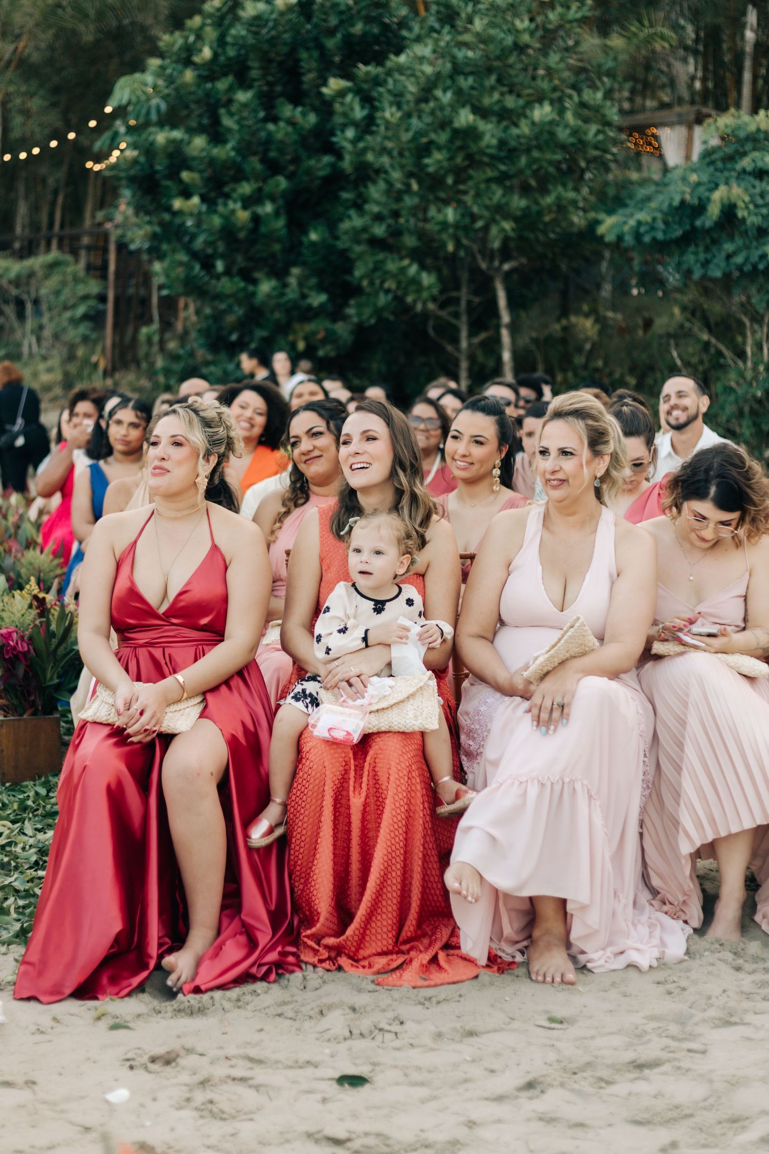 Bridesmaids in pink and red dresses sitting together on a beach for a group photo.