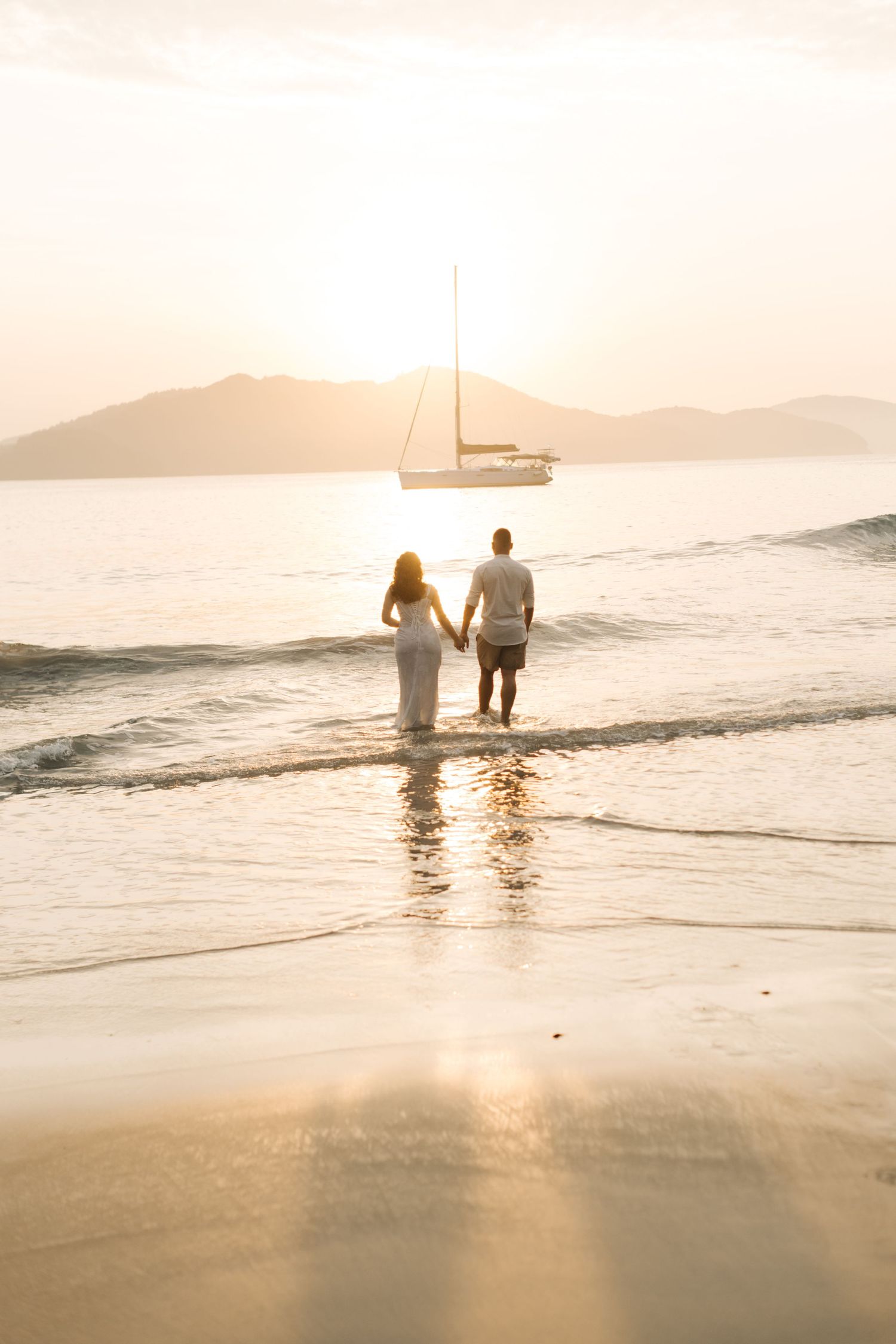 A couple wades in ocean water at sunset with a sailboat anchored in the distance.