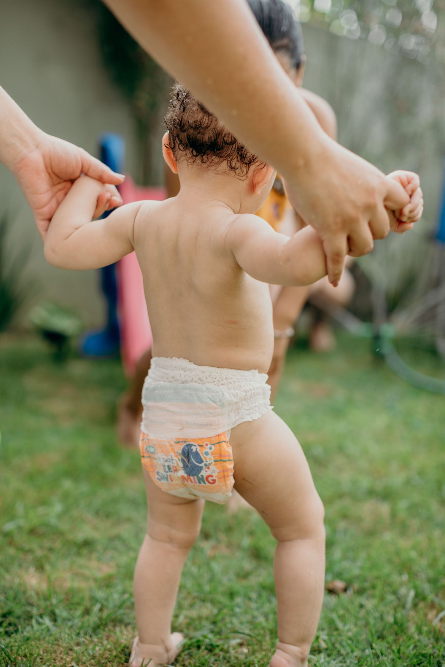 Close-up of tiny feet and hands being guided by adults on green grass.