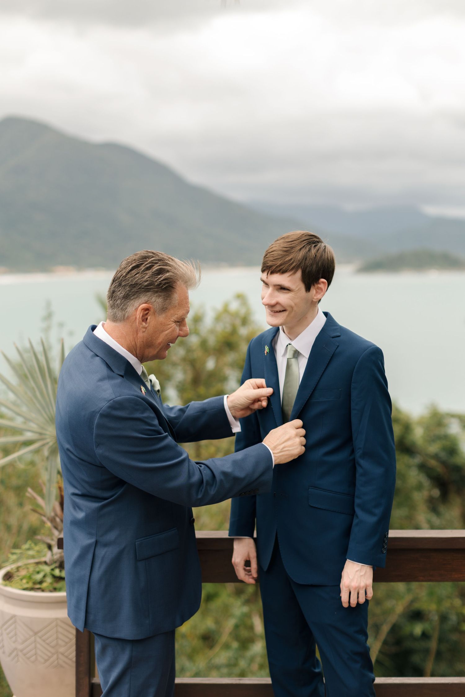 Two people in navy suits share a lighthearted moment on an outdoor deck with mountains and water in the background.