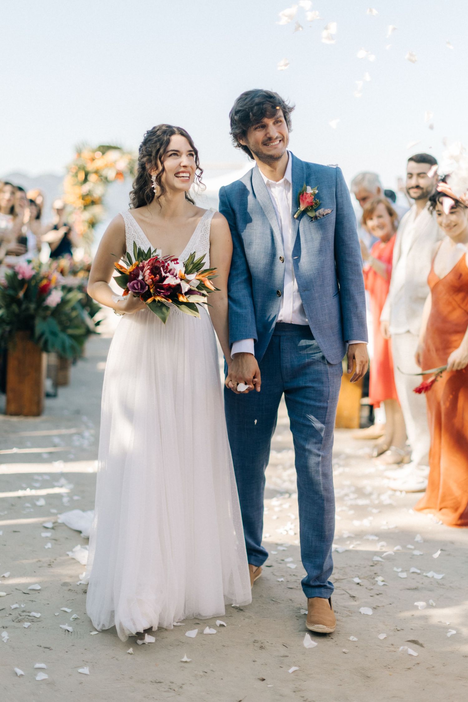 Newlyweds share a joyful moment walking together on a sunlit beach path with scattered petals.