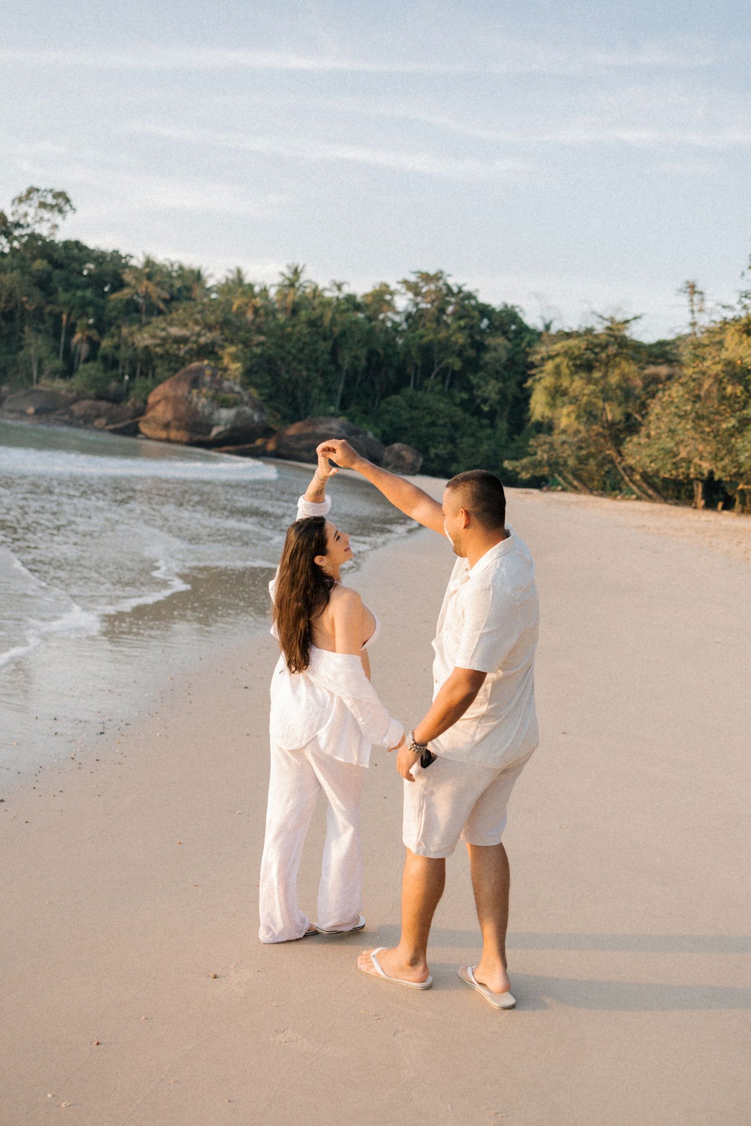 Couple in white clothing dancing together on an empty beach surrounded by lush greenery.
