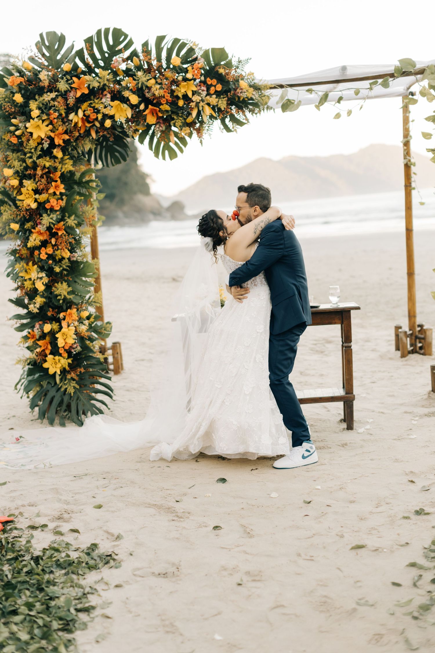 Newlyweds share romantic embrace under tropical flower arch on beach during sunset ceremony.