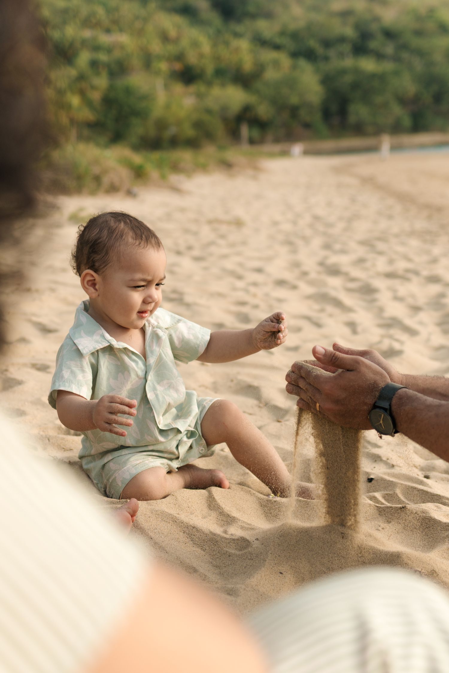 A small child plays with sand on a sunny beach while someone helps build a sandcastle.