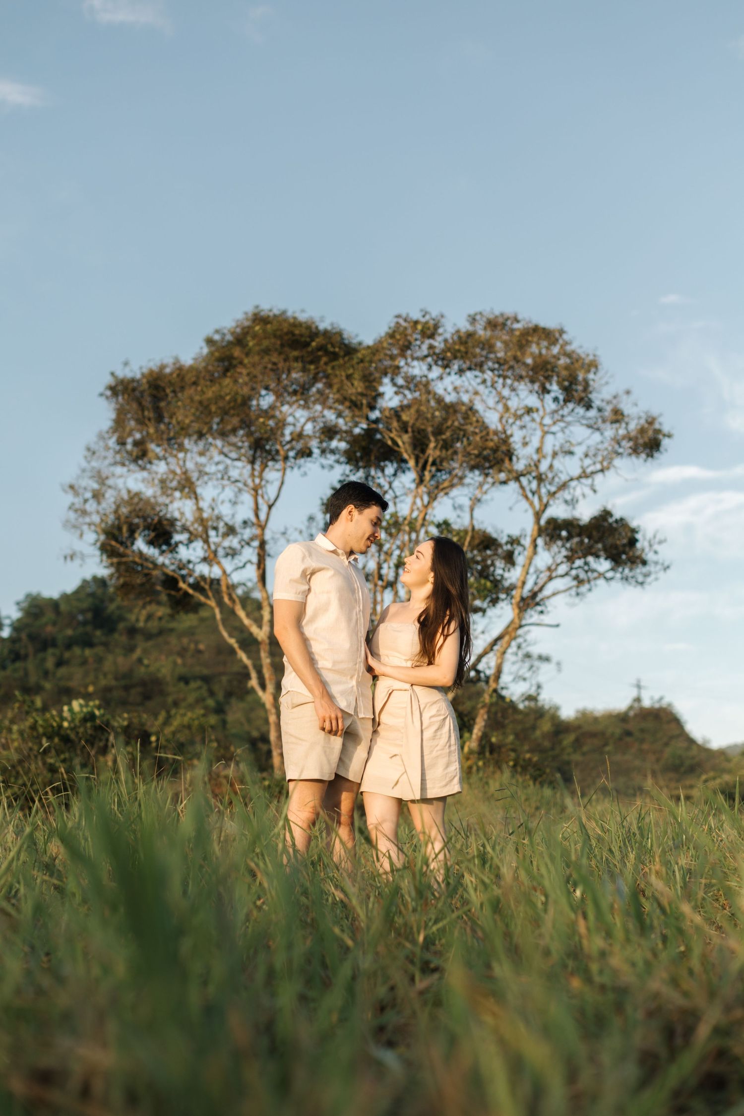 Couple in coordinating beige outfits standing together in tall grass with trees behind them.