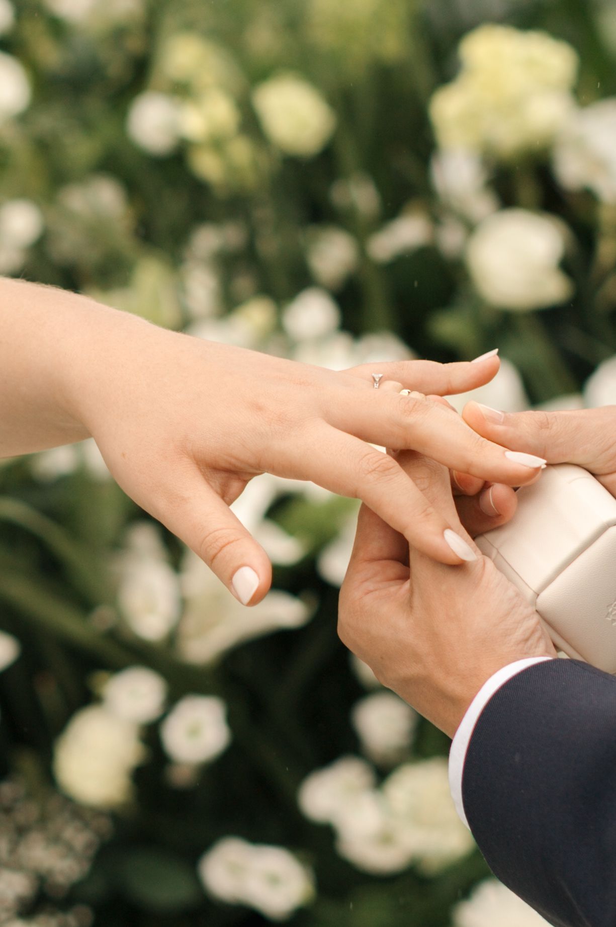Close-up of hands during wedding ring exchange against white floral backdrop.