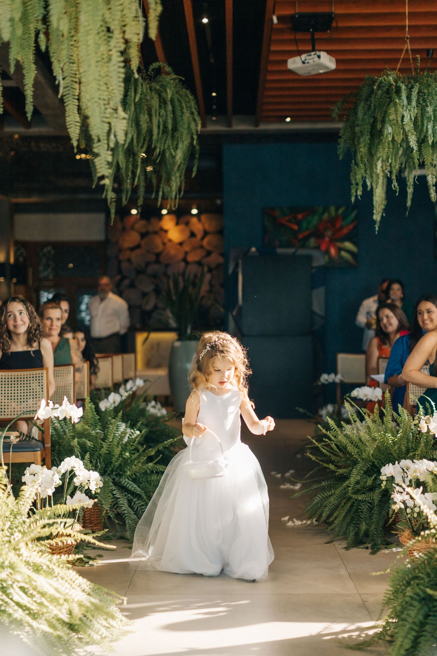 A flower girl in white dress twirls down an aisle decorated with ferns and hanging greenery.
