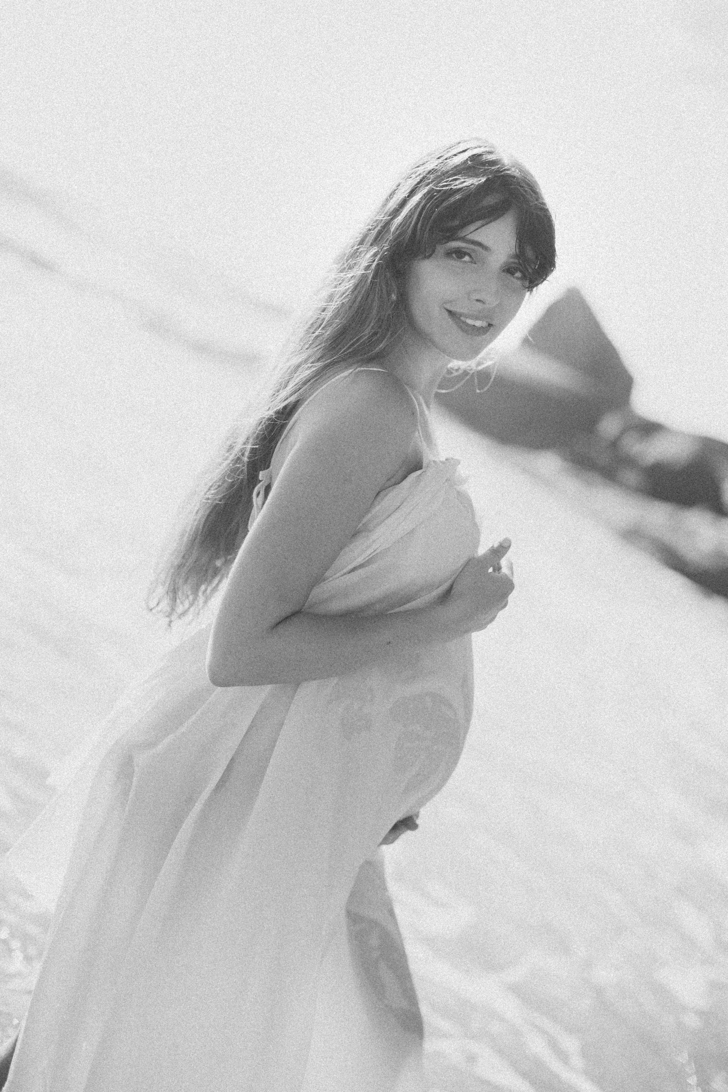 A dreamy black and white portrait of someone by the ocean in a flowing white dress with long dark hair.