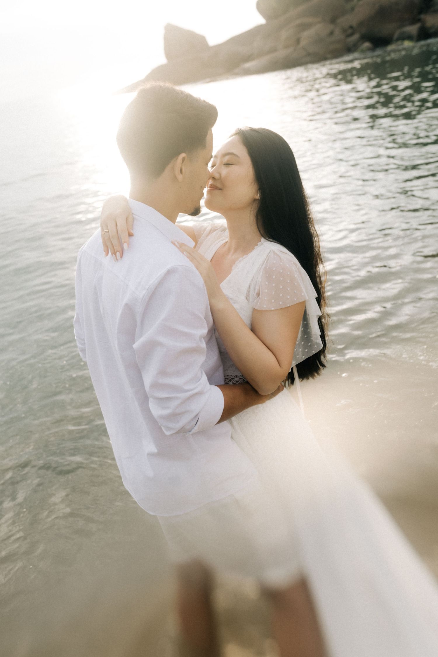 Tender beach moment between lovers wearing white as sunlight sparkles on the water.