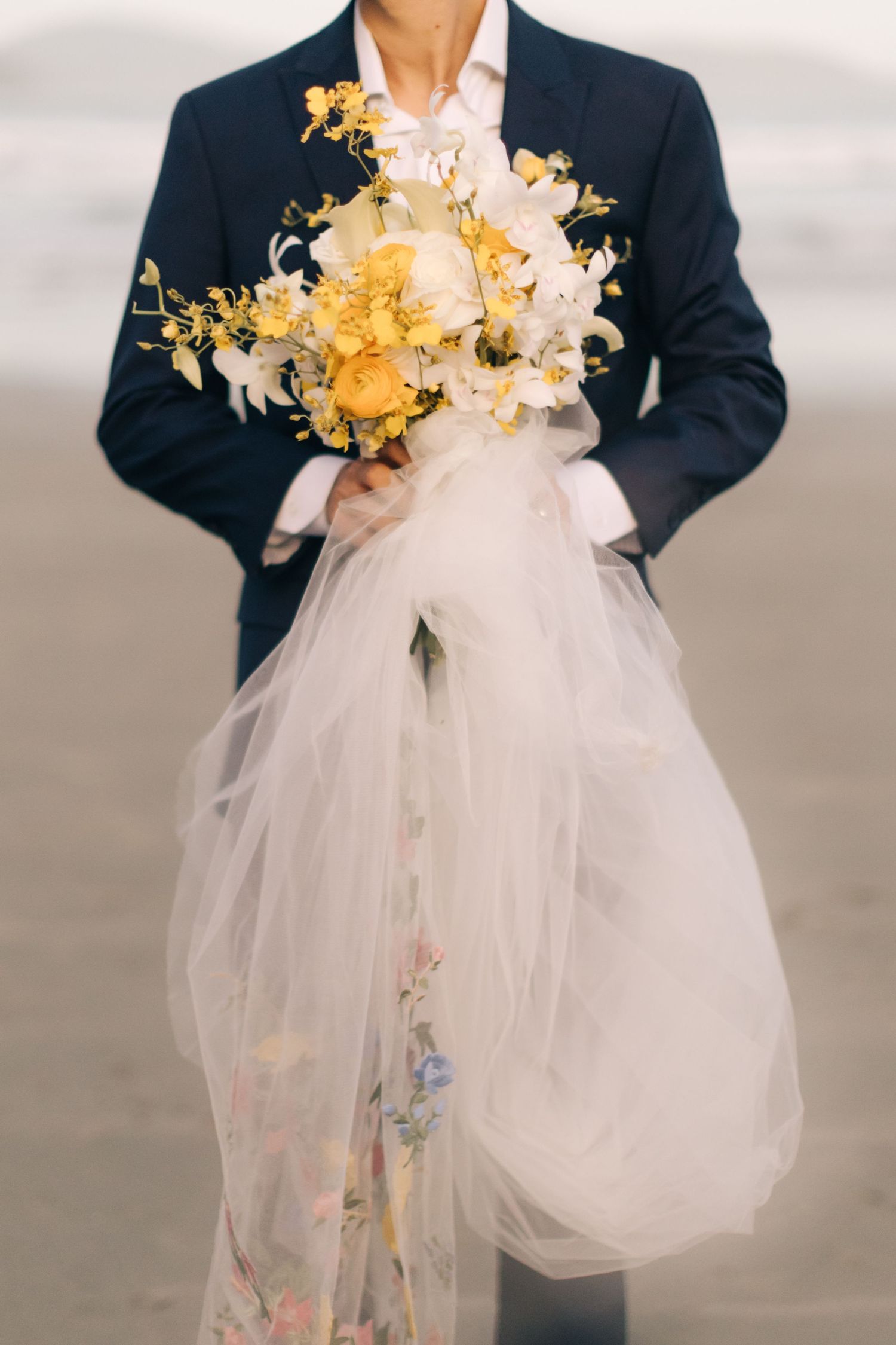 A beautiful yellow and white floral bouquet held against a navy suit with flowing white tulle.