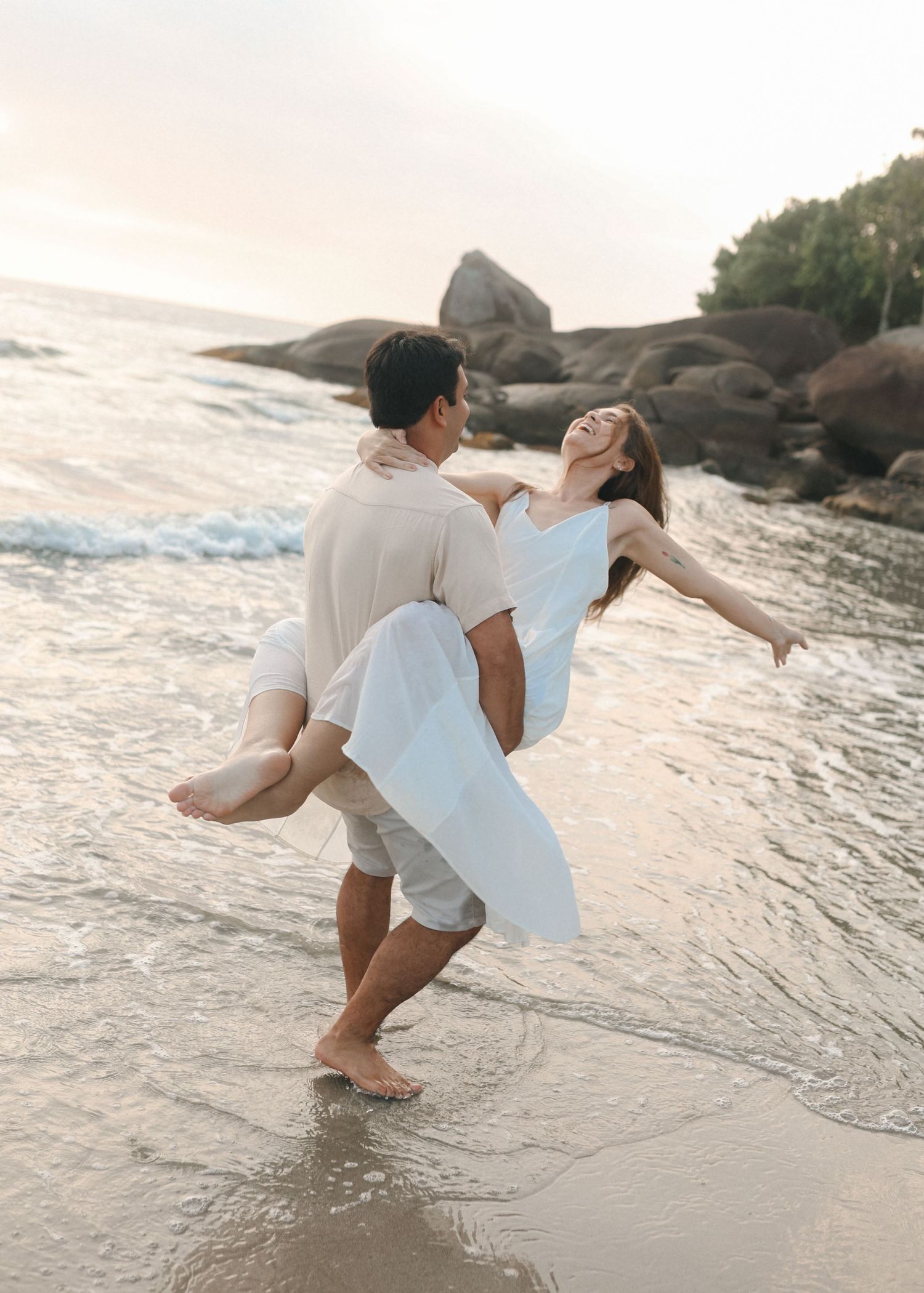 A romantic beach scene with someone being carried while waves crash at their feet.