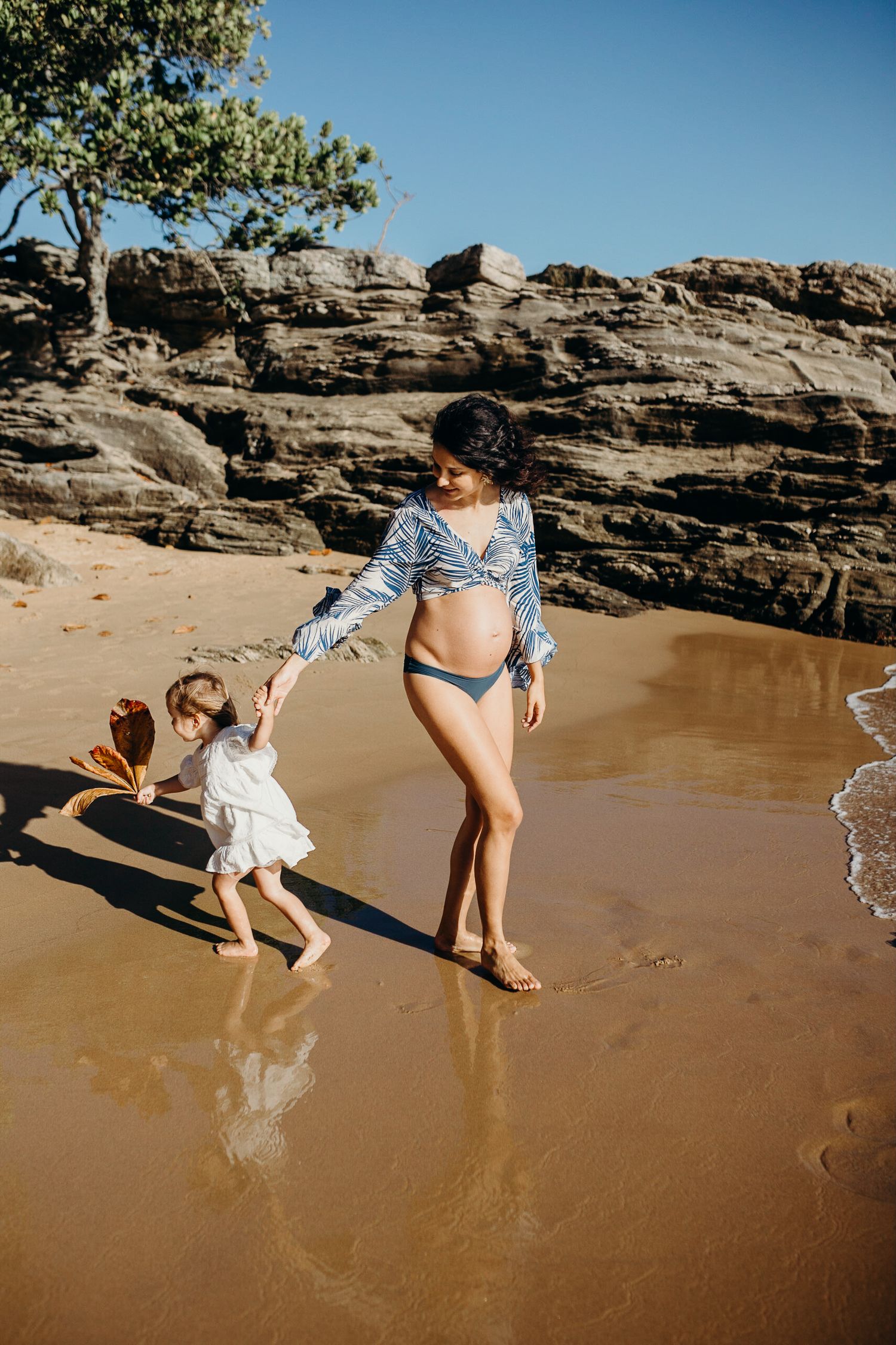 Mother and child play together at the edge of ocean waves near rocky cliffs.