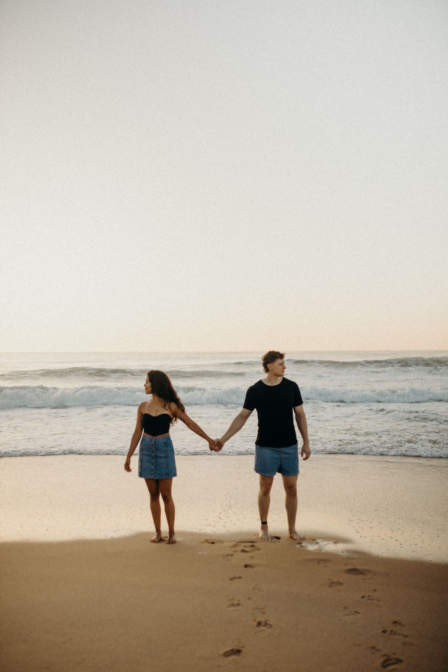 Couple holding hands while standing barefoot on beach with waves and sunset in background.