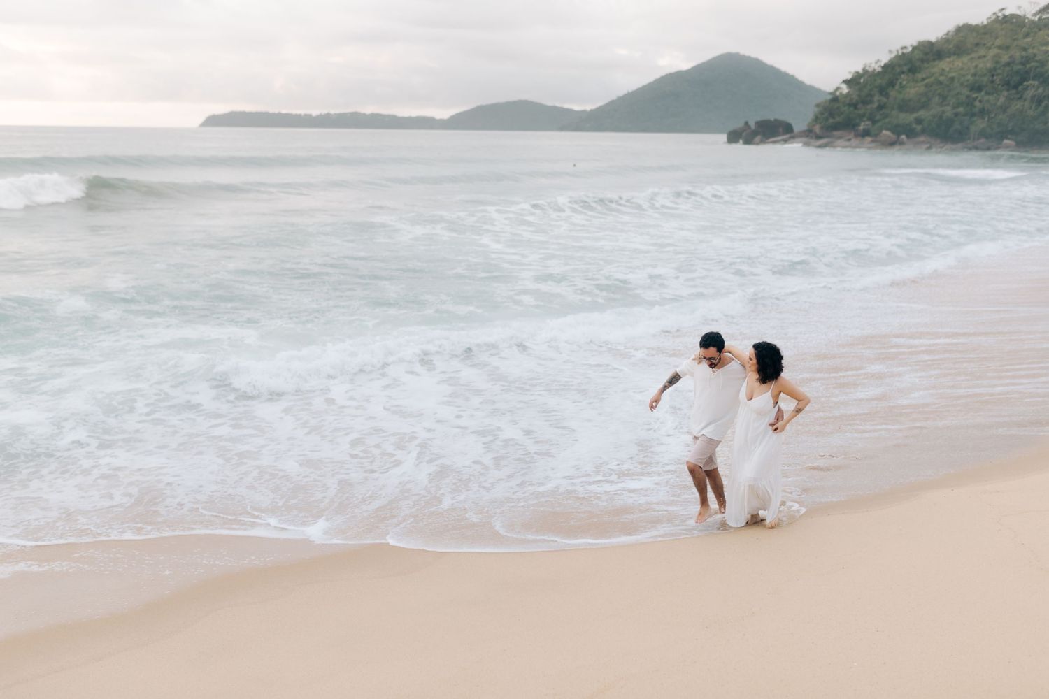 A couple walks along a scenic beach at sunset with mountains in the background.