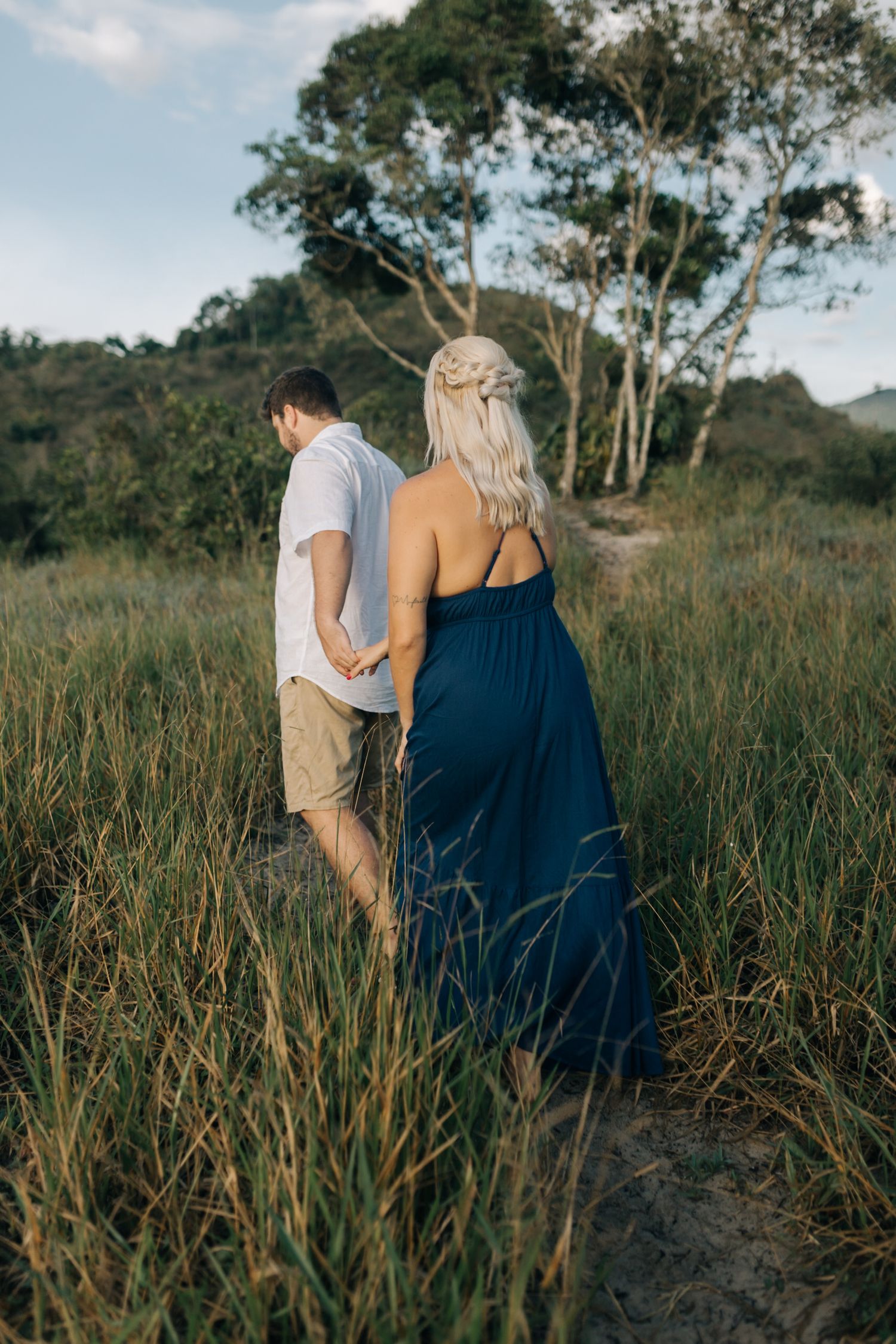 Intimate couple walking through wild grass field with eucalyptus trees in background.