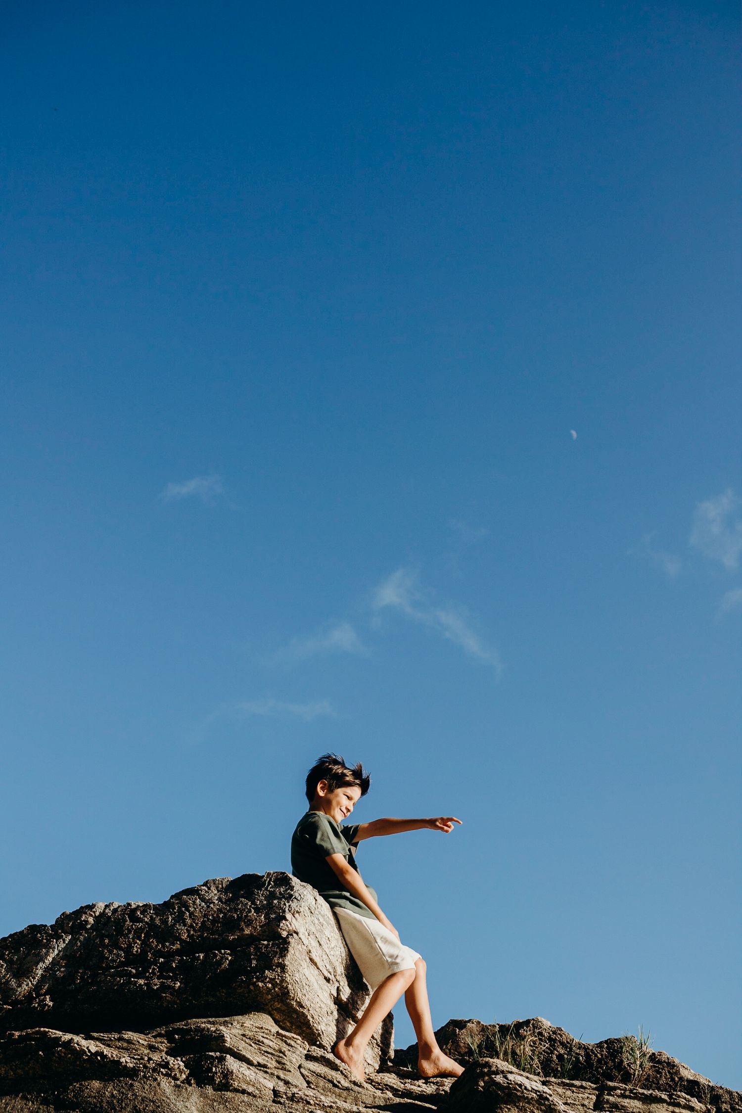 Figure sits contemplatively on rocky outcrop overlooking ocean under blue sky.