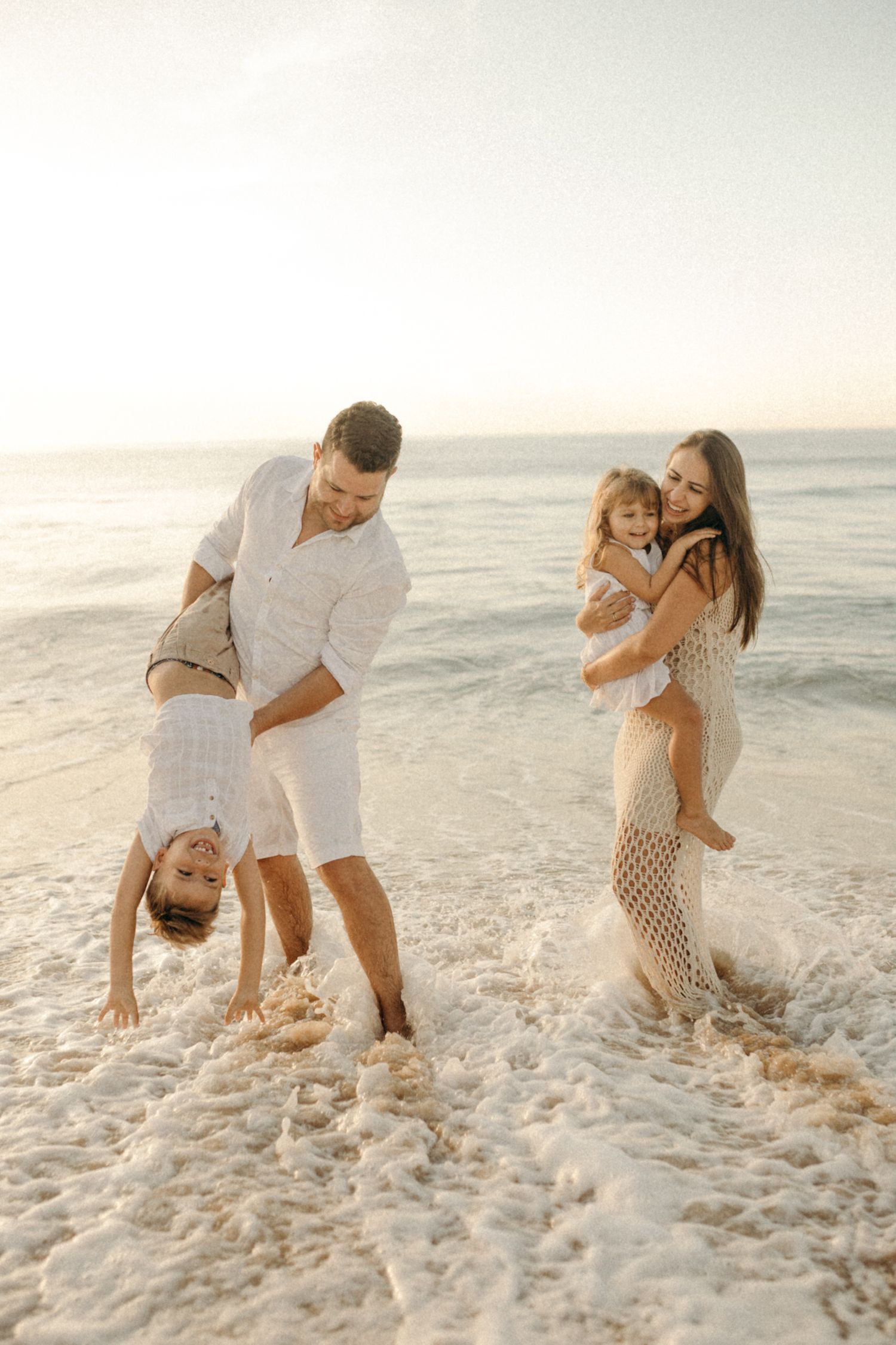 Family playing in ocean waves on sandy beach during golden hour sunset.