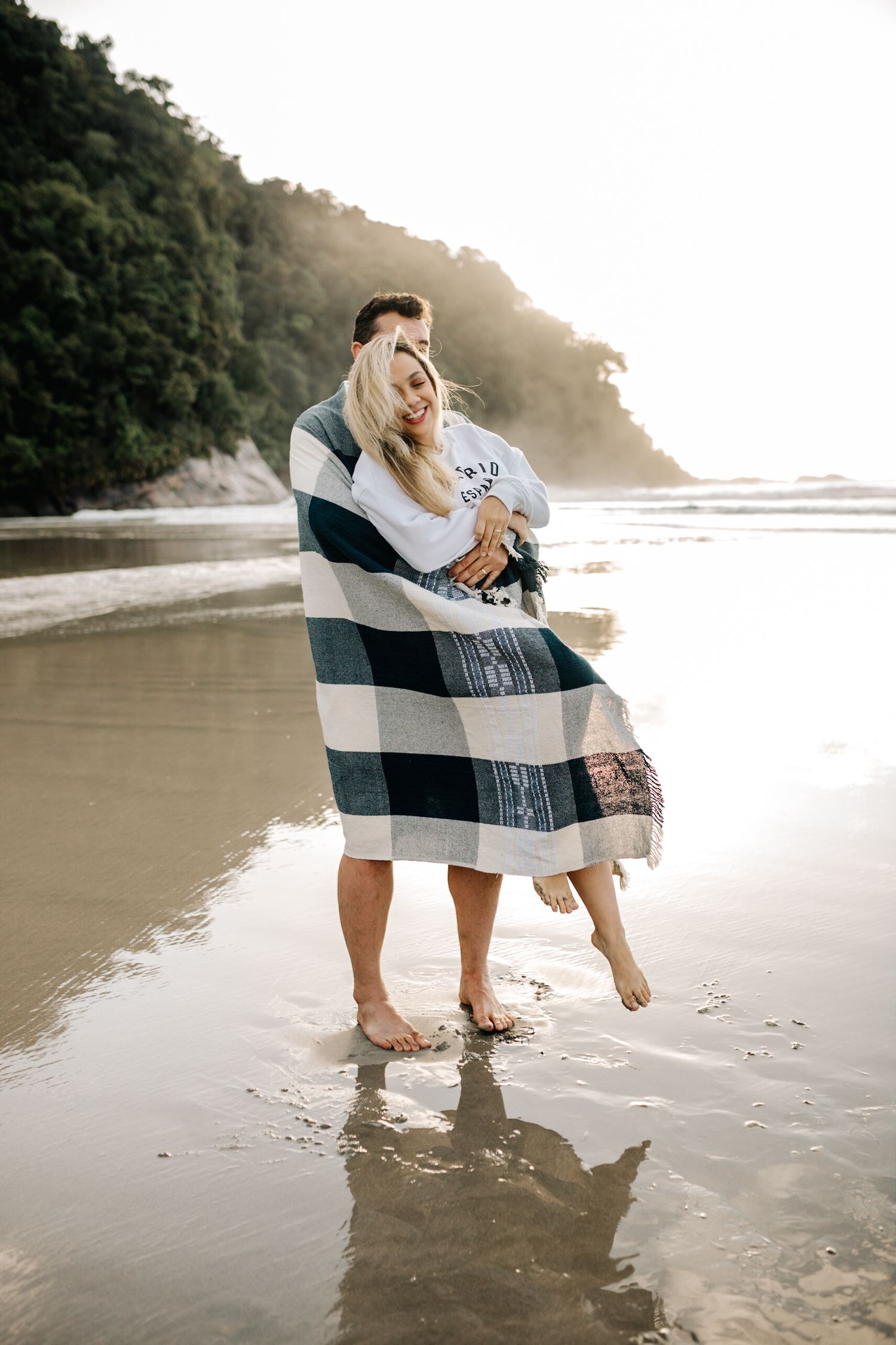A couple wrapped in a blue plaid blanket embraces on a peaceful beach at dusk.