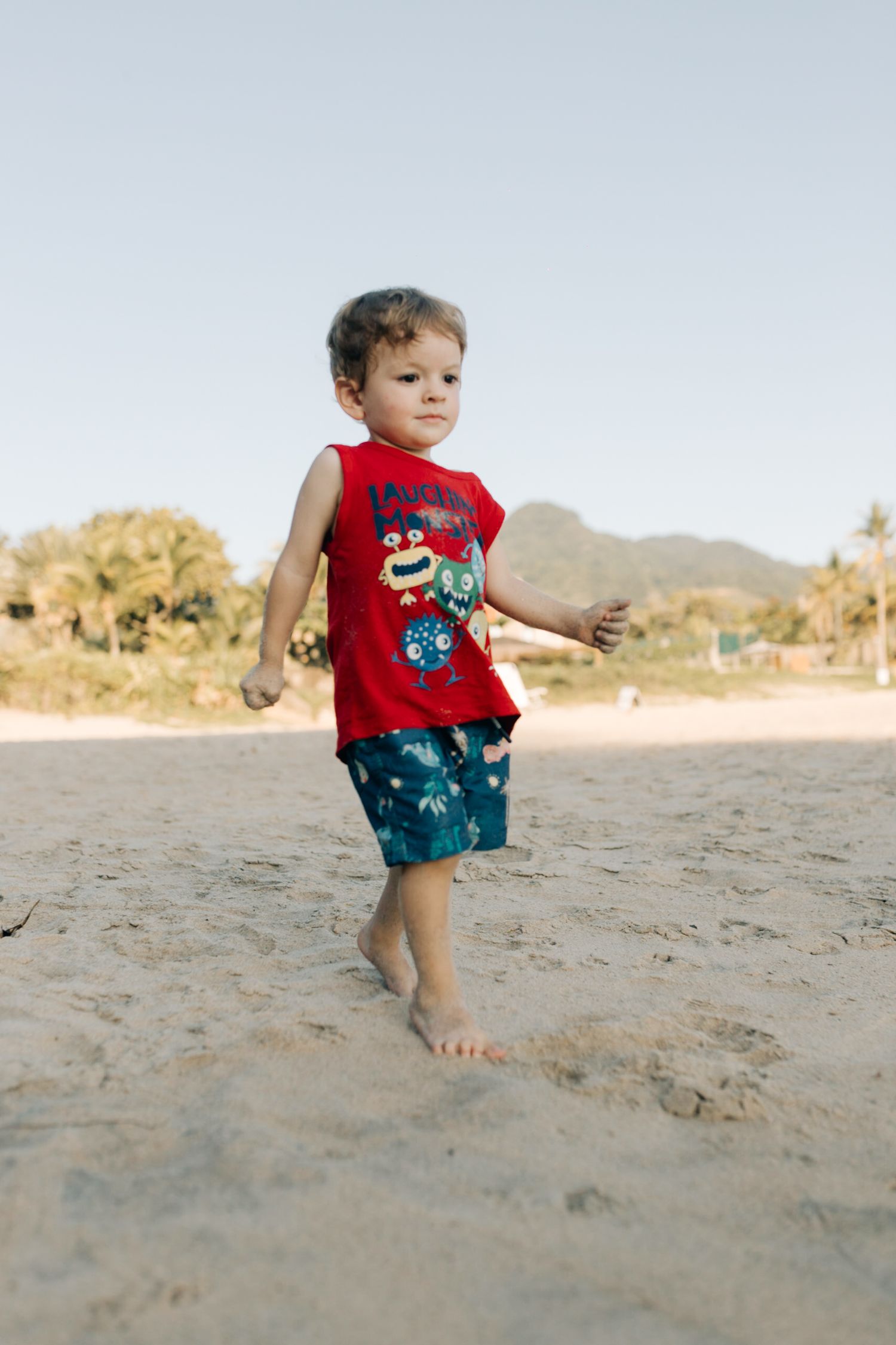 Small child wearing red shirt and blue shorts playing on sandy beach with mountains in background.