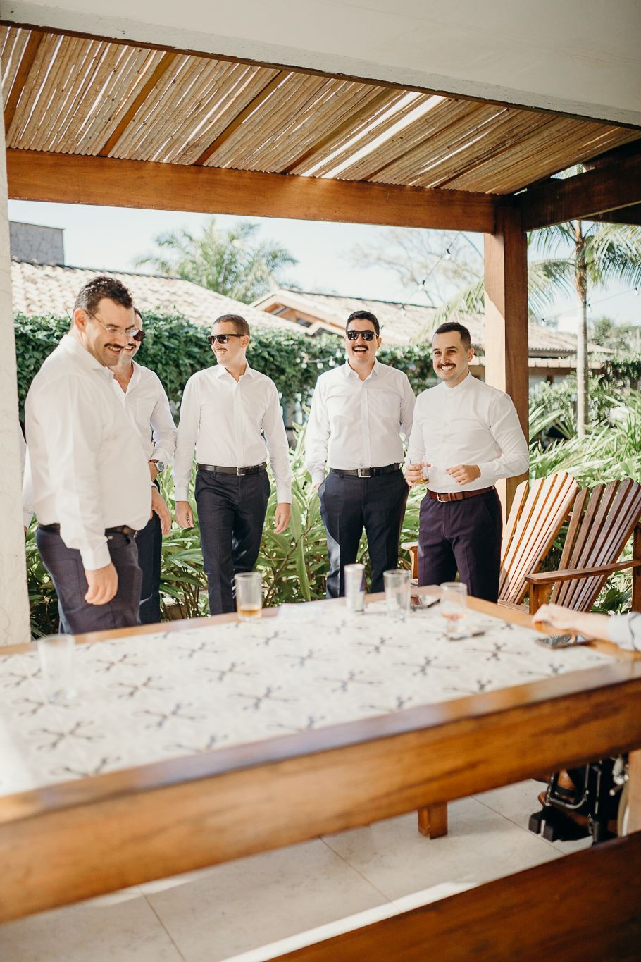 Wedding party in white shirts gather under wooden pergola at outdoor venue.