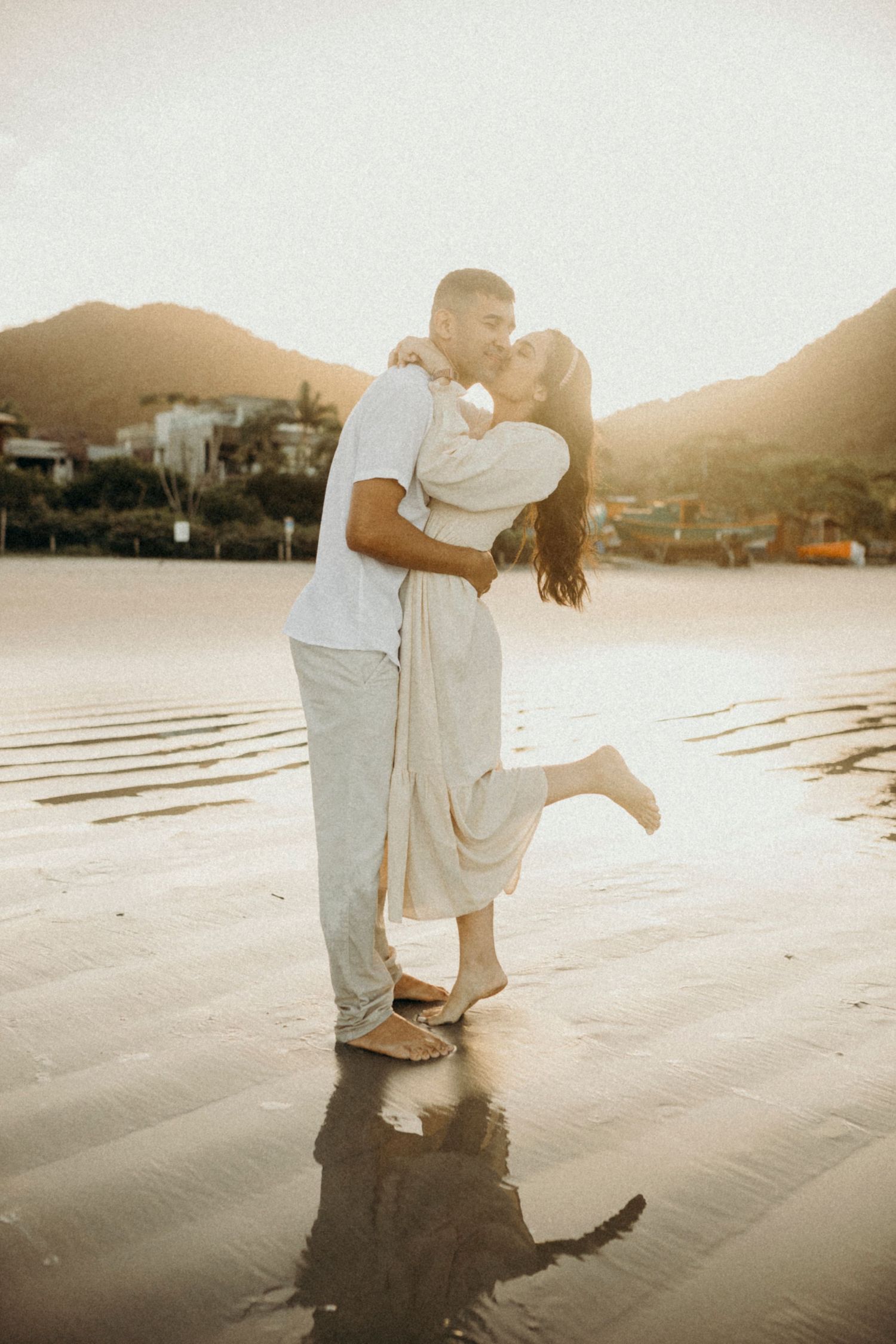 A romantic couple embraces on a beach at sunset with mountains in the background.