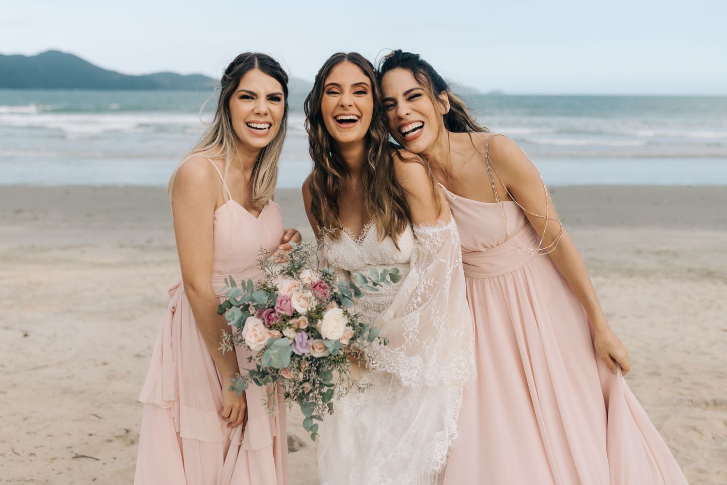 Three women in pink bridesmaid dresses and wedding gown pose joyfully on a beach during sunset.