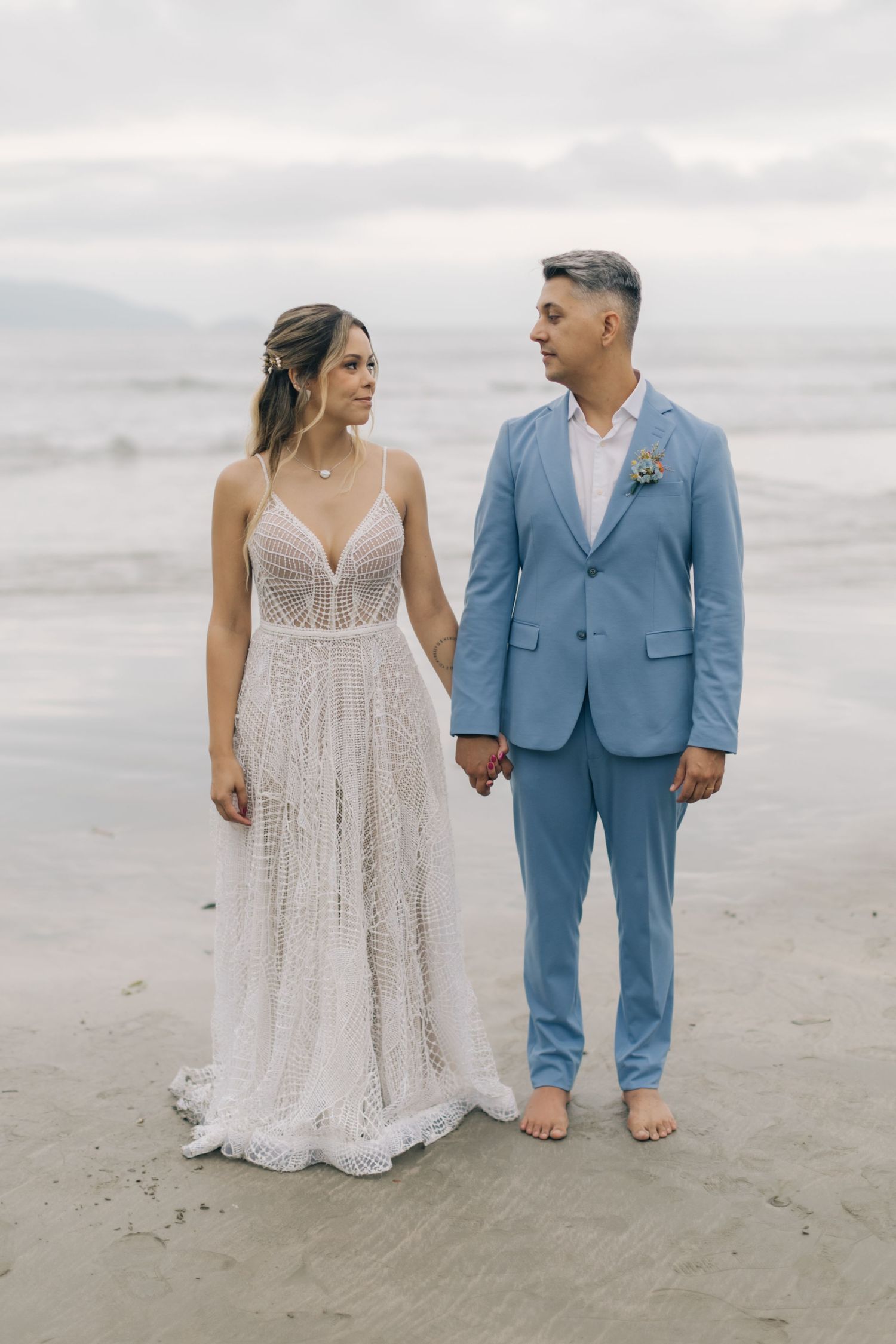 A couple walks barefoot on the beach, with the bride in lace and groom in light blue.