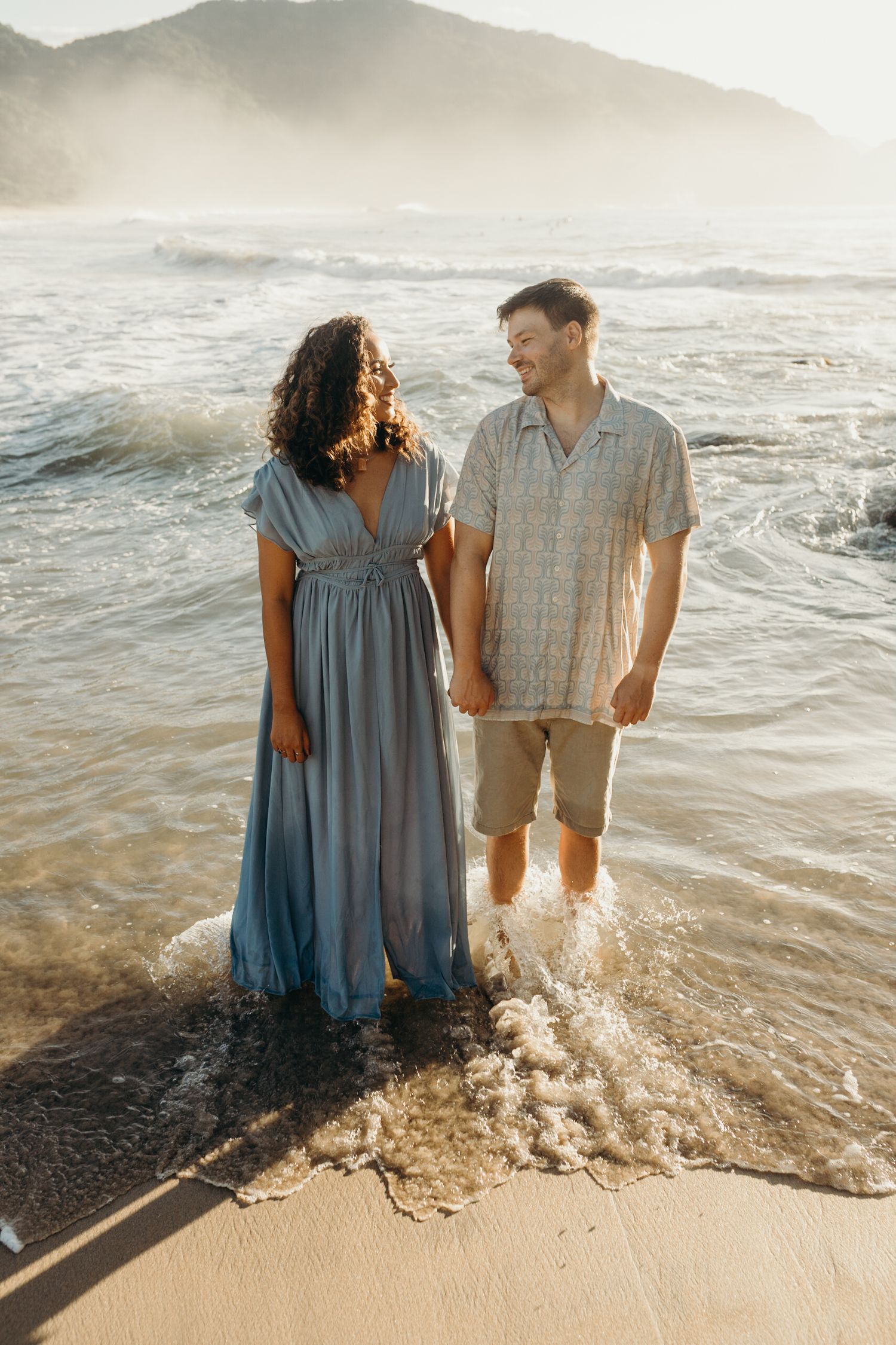 Couple standing in ocean waves at sunset on a sandy beach.