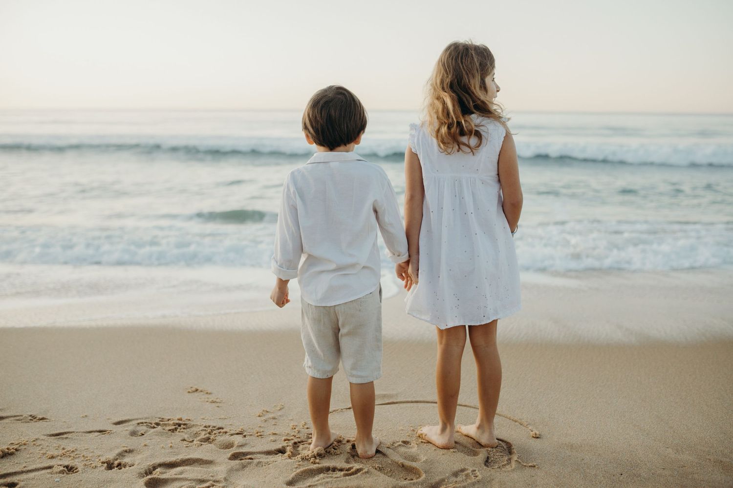 Two children walking along the shoreline in white outfits as waves roll in at sunset.