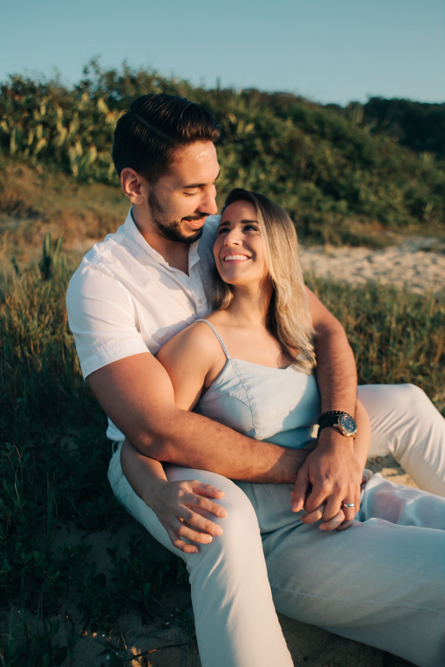 Romantic couple in white outfits sharing an intimate moment on a hillside during golden hour.