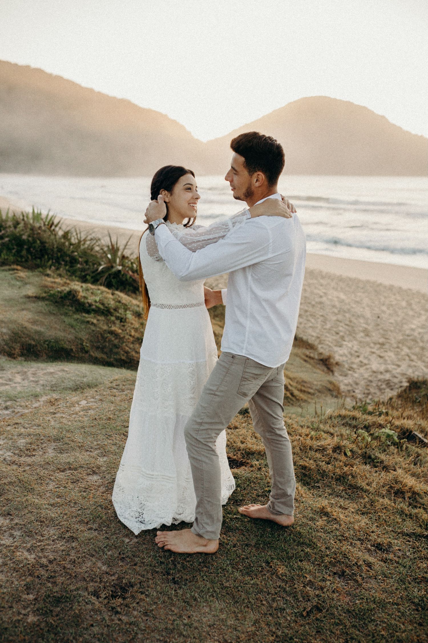 A couple shares a tender moment on a grassy cliff overlooking a misty beach at sunset.