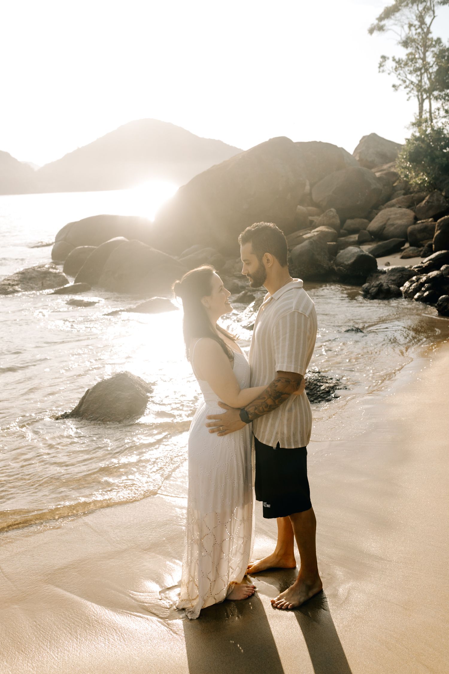 Romantic beach photoshoot featuring couple embracing by rocky coastline at sunset.