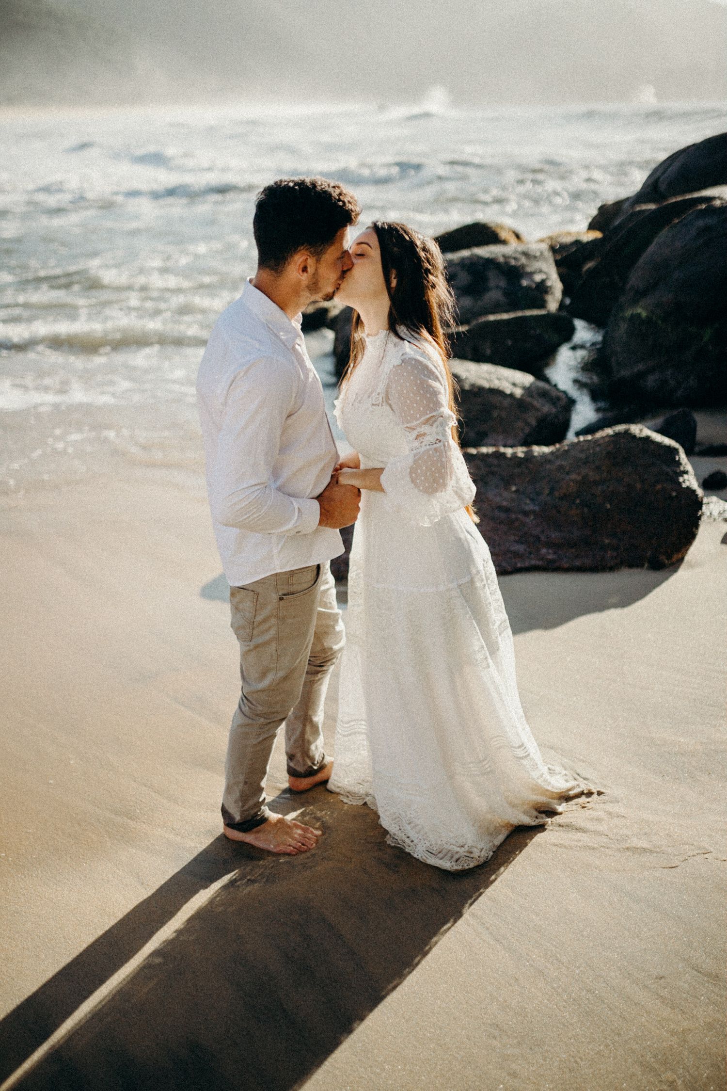 A couple shares a romantic moment on a beach with dramatic rocky coastline in the background.