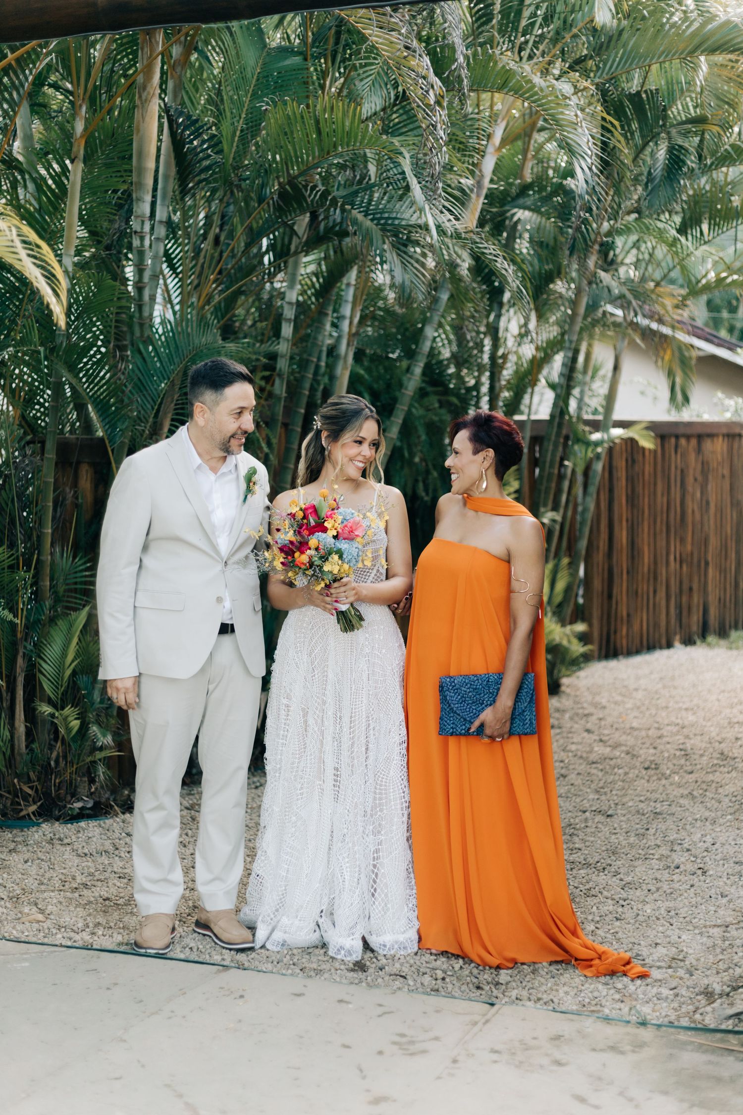 Wedding party in white and orange attire poses together on tropical path lined with palm trees.