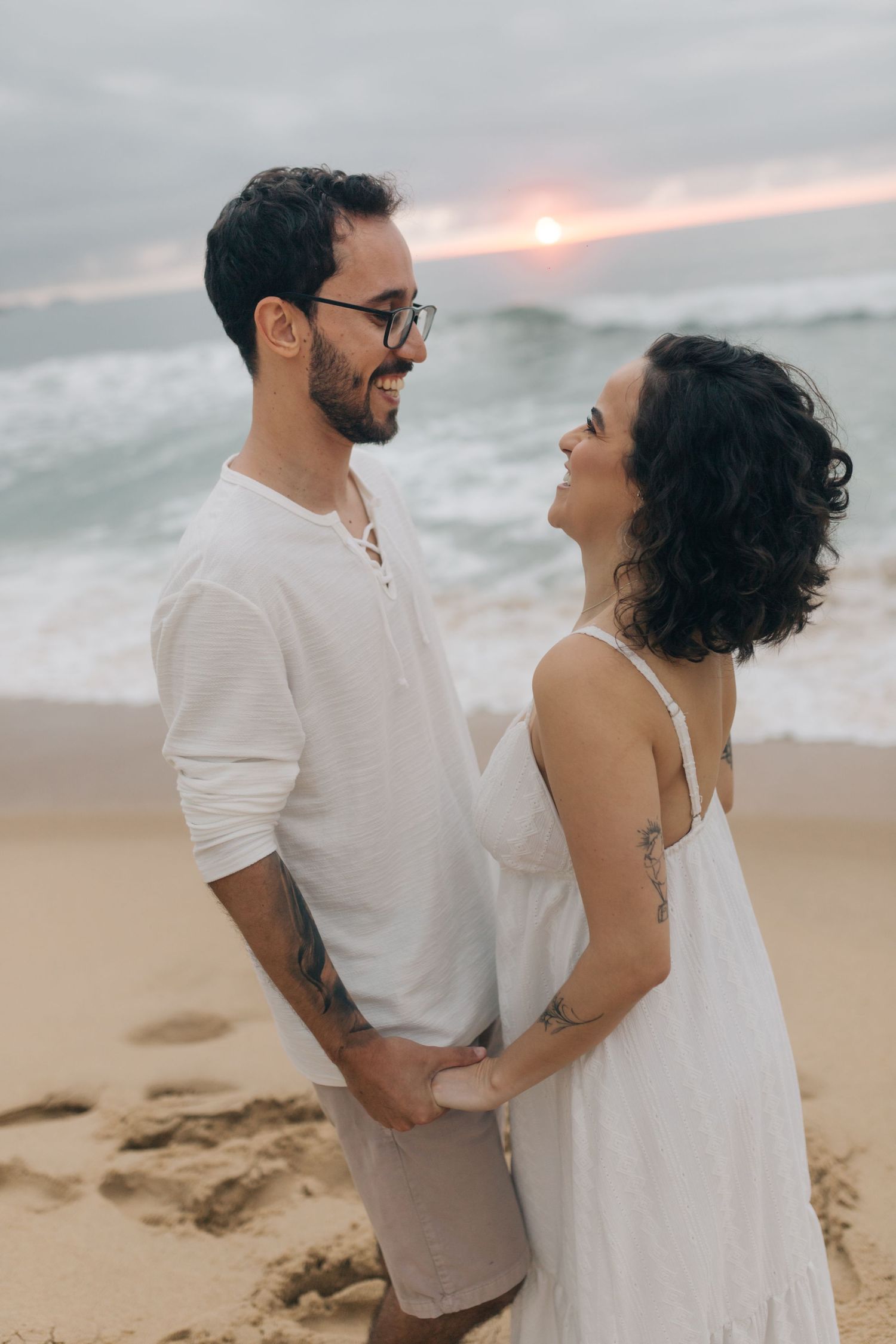 A couple in white outfits laughs together on a beach with waves crashing behind them.