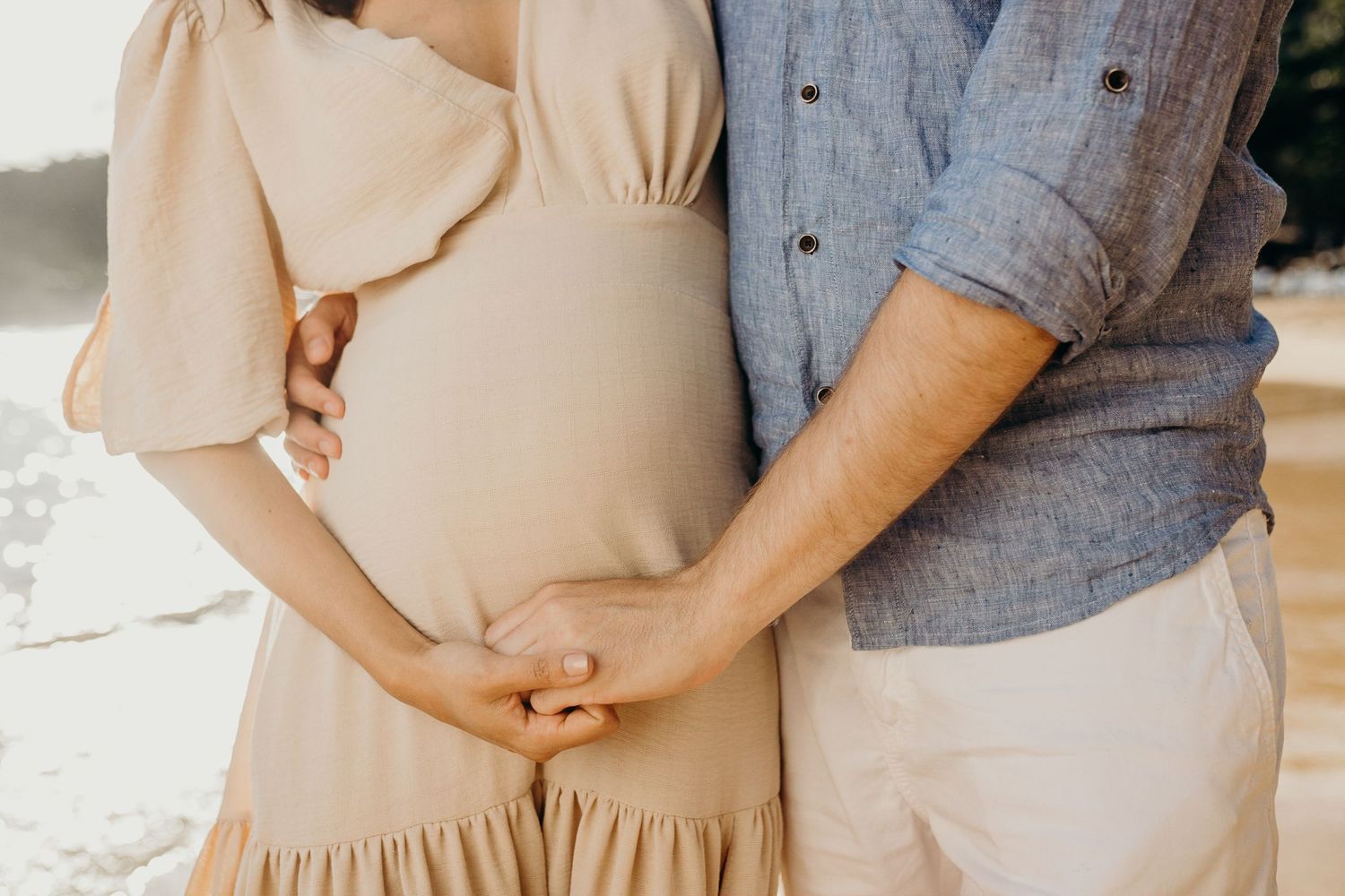 Close-up shot showing hands cradling a baby bump on a sunlit beach.