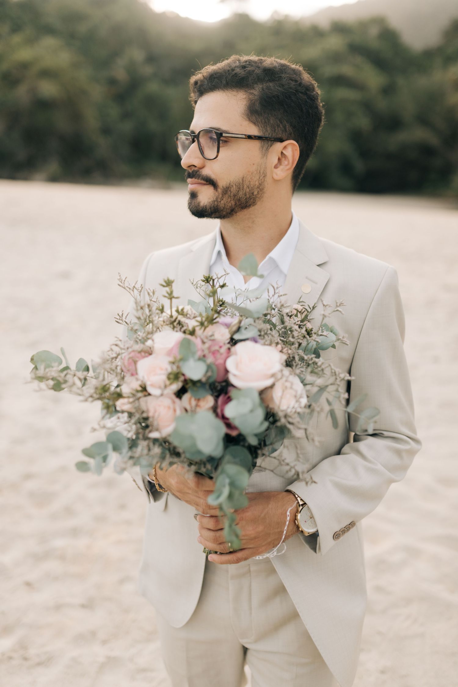 A person in a beige suit holds a delicate bouquet of pink and white flowers with eucalyptus.