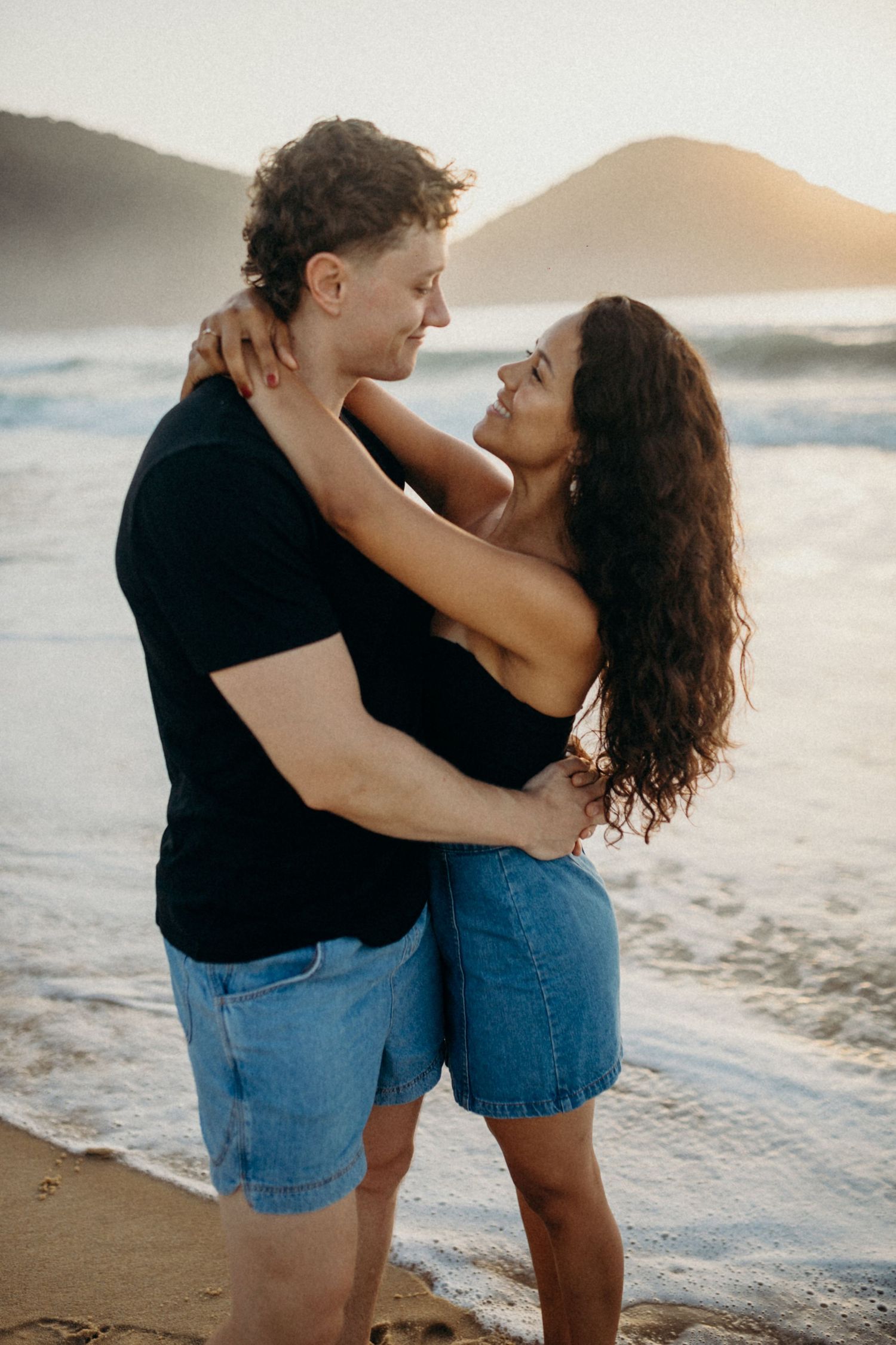 Romantic beach embrace with mountains and sunset waves creating a dreamy coastal backdrop.