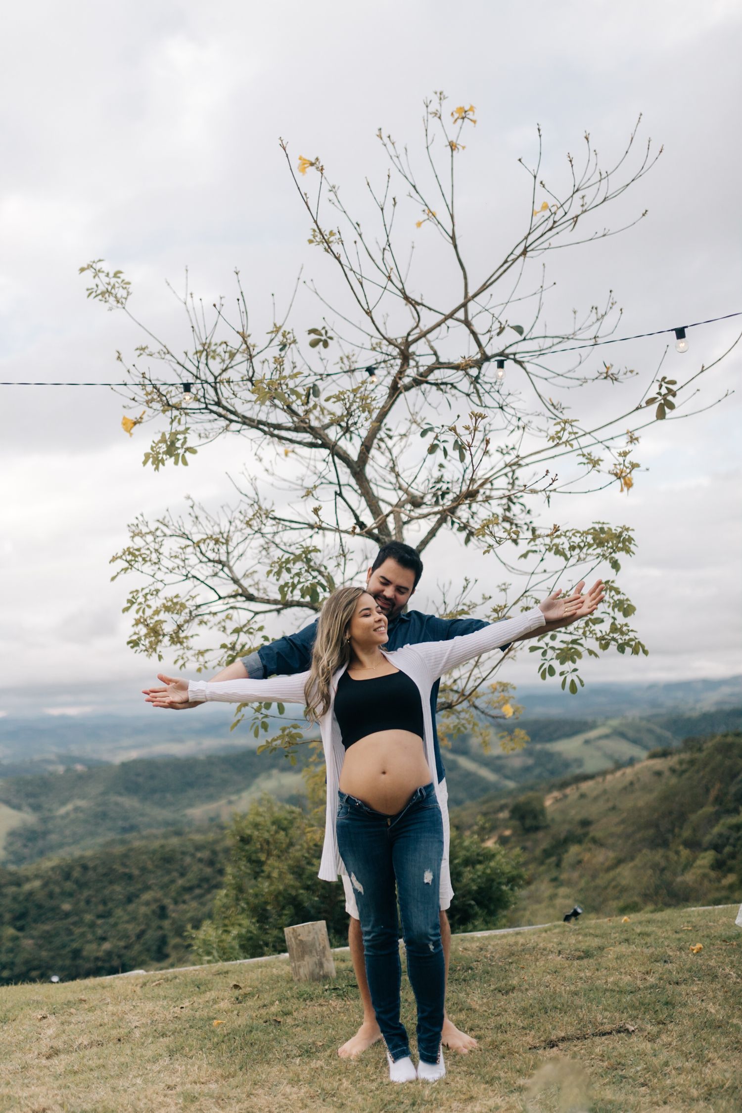 A couple poses playfully under a bare tree on a hilltop with mountain views in the background.