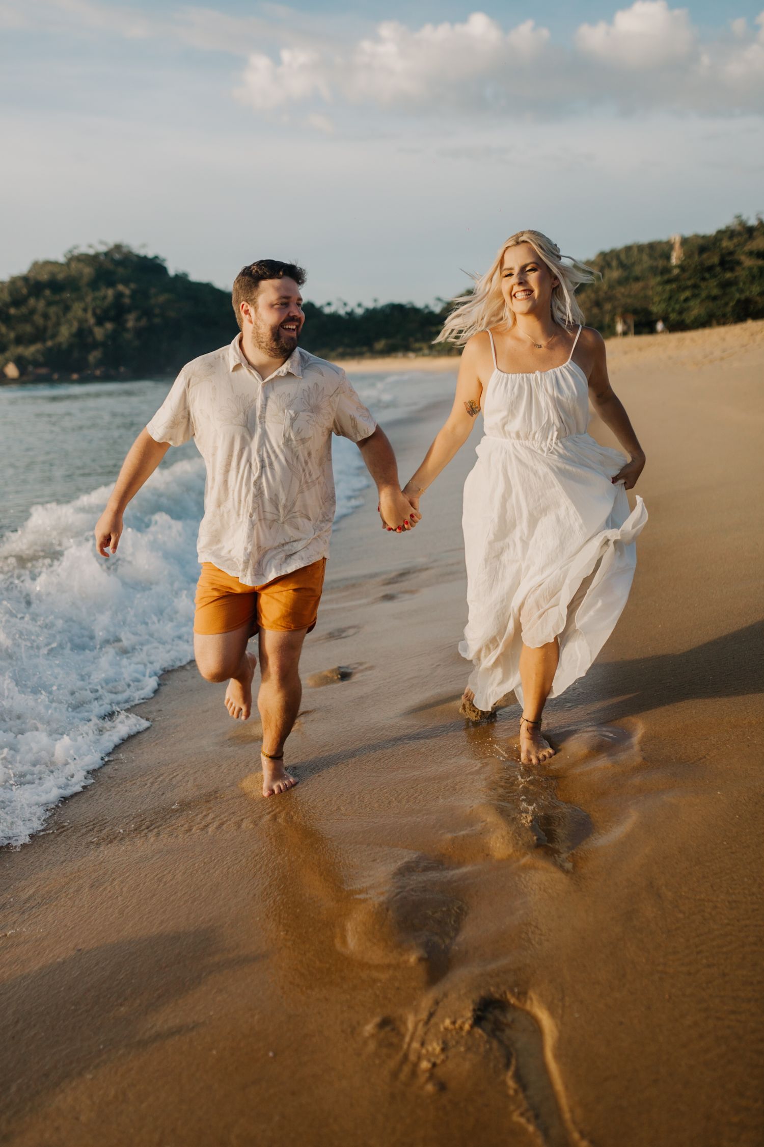 Energetic couple running through shallow waves on tropical beach at sunset.