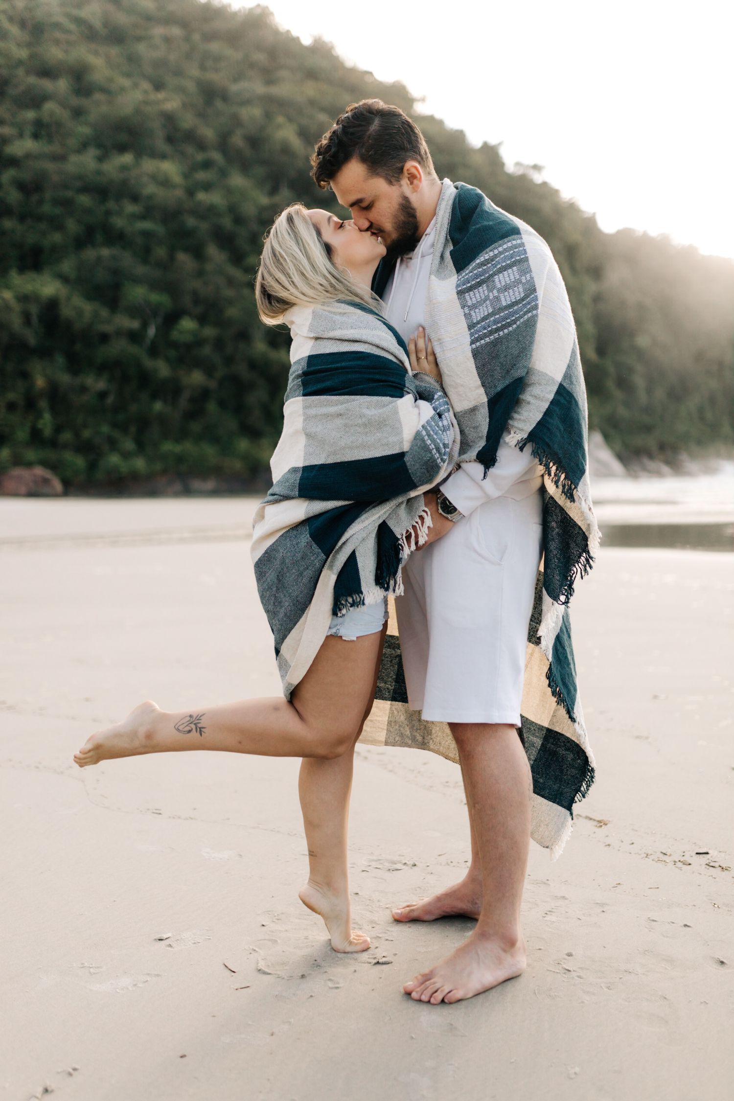 A couple shares a romantic kiss while wrapped in a plaid blanket on a sandy beach.